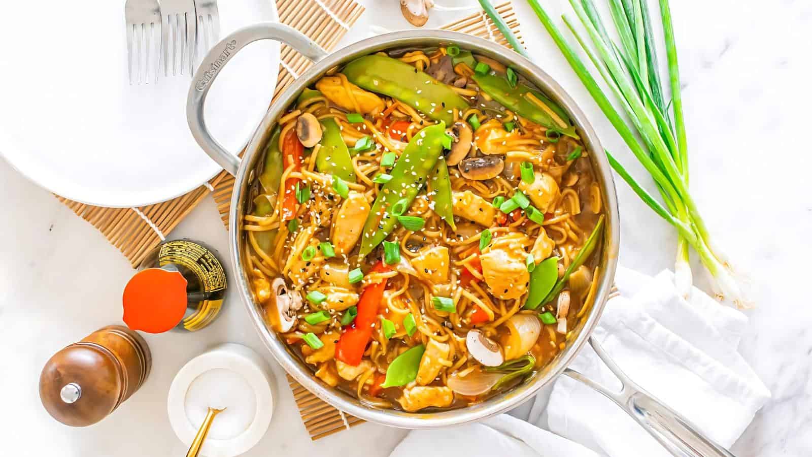 A colorful stir-fry with noodles, vegetables, and sesame seeds in a pan. Chopsticks, a pepper grinder, and green onions are nearby on a white surface.