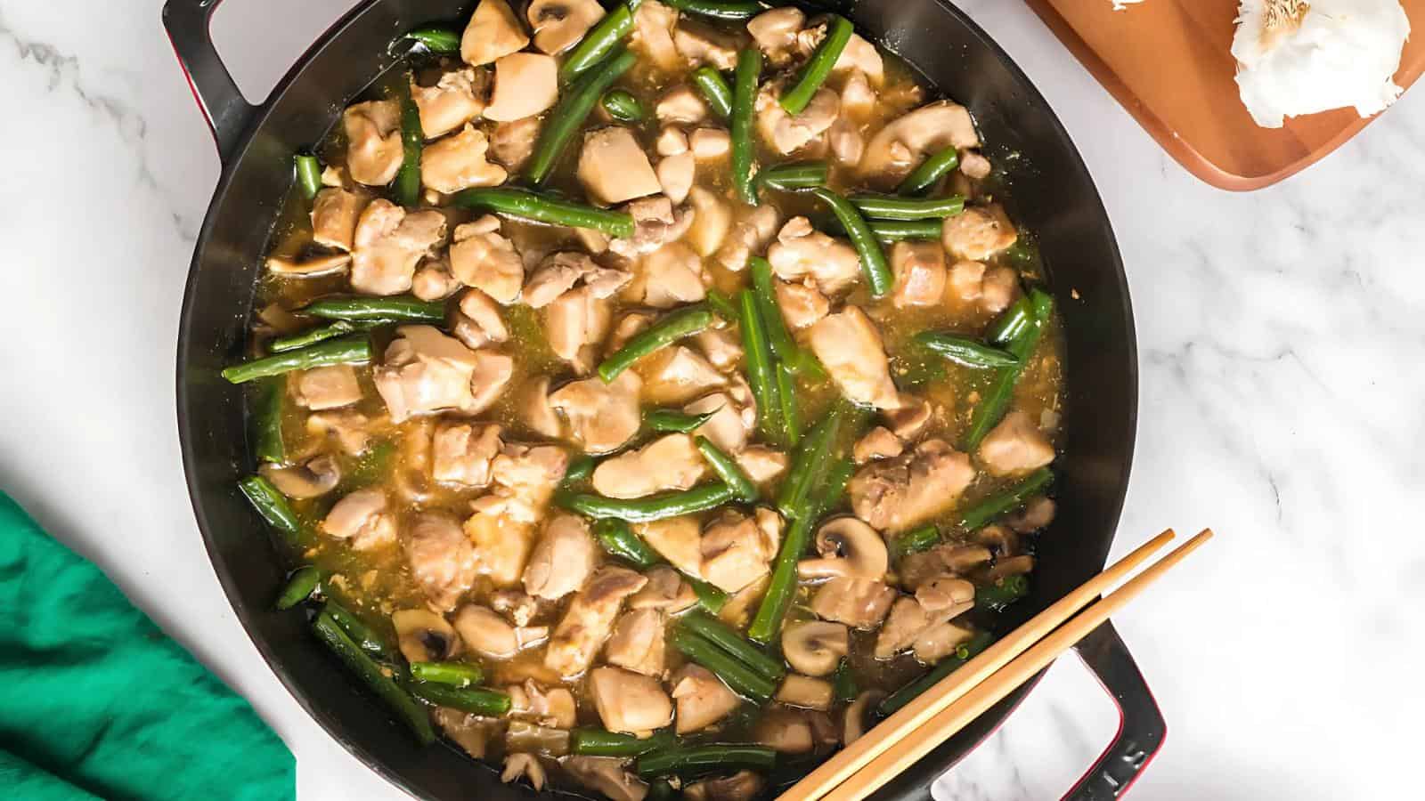 A pan filled with cooked chicken pieces, green beans, and mushrooms in a brown sauce, with chopsticks resting on the side.