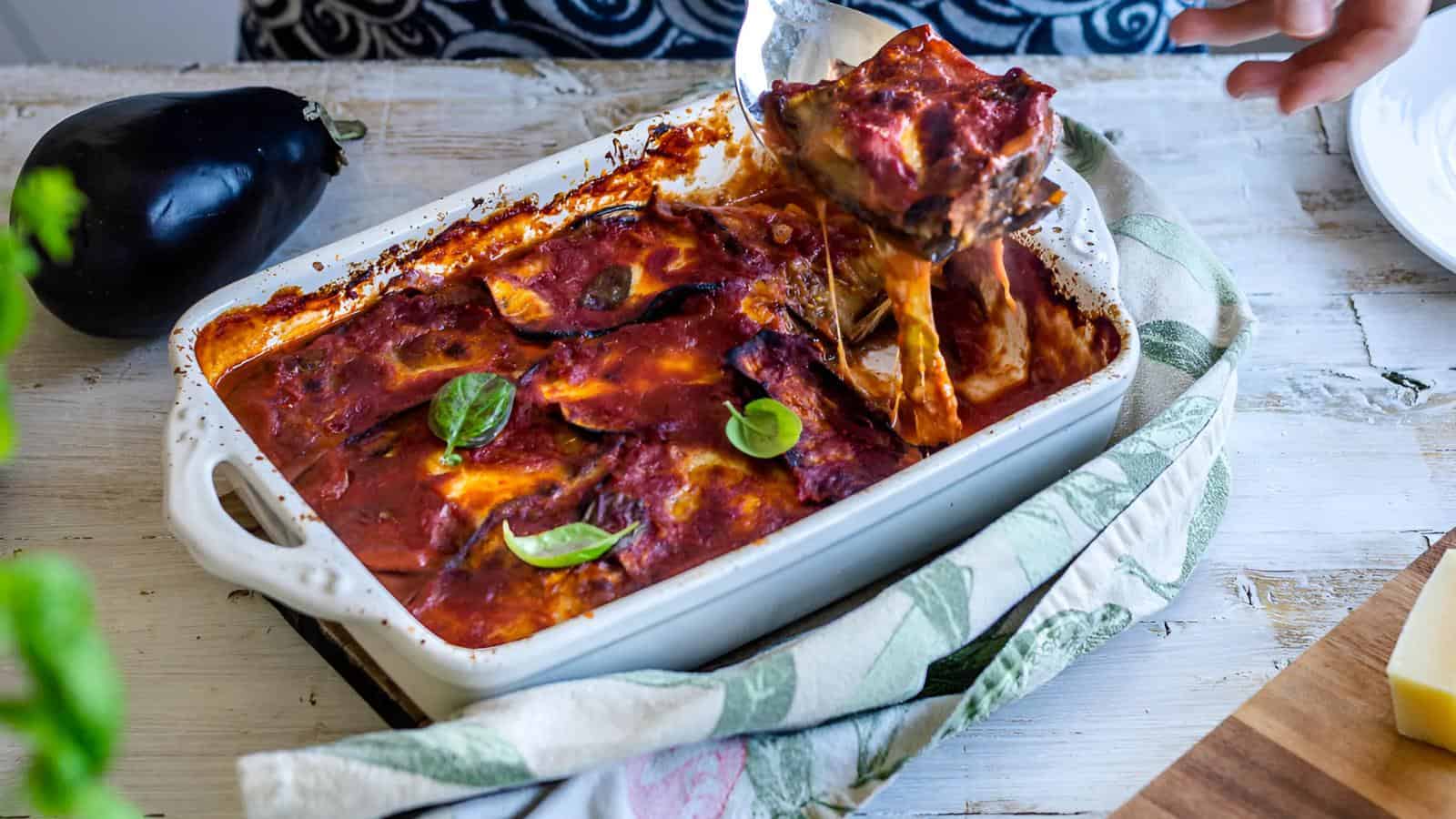 A person serves a slice of baked eggplant parmesan from a casserole dish, with fresh basil leaves on top. An eggplant is on the table nearby.