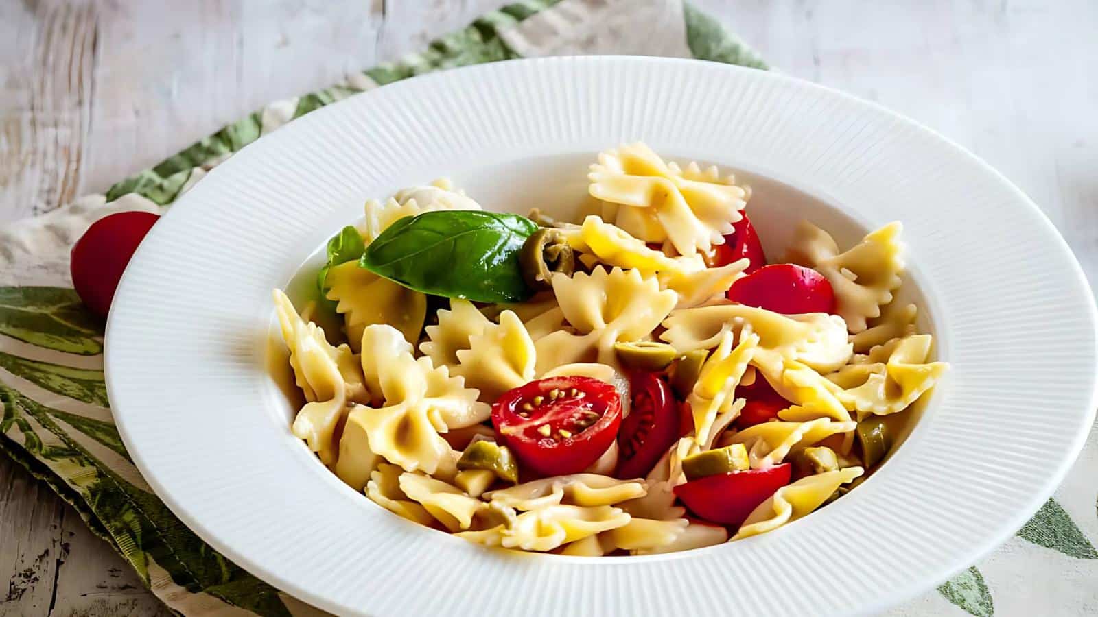 A white bowl of bowtie pasta with cherry tomatoes, green olives, and a basil leaf, placed on a green and white cloth.