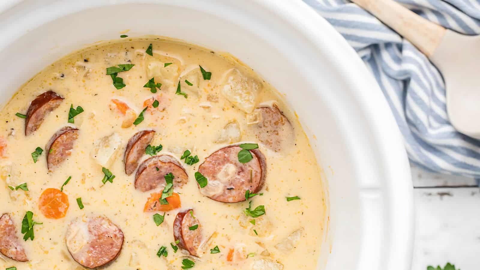 A creamy soup with sliced sausages, potatoes, carrots, and herbs in a white bowl. A striped cloth napkin is partially visible in the background.