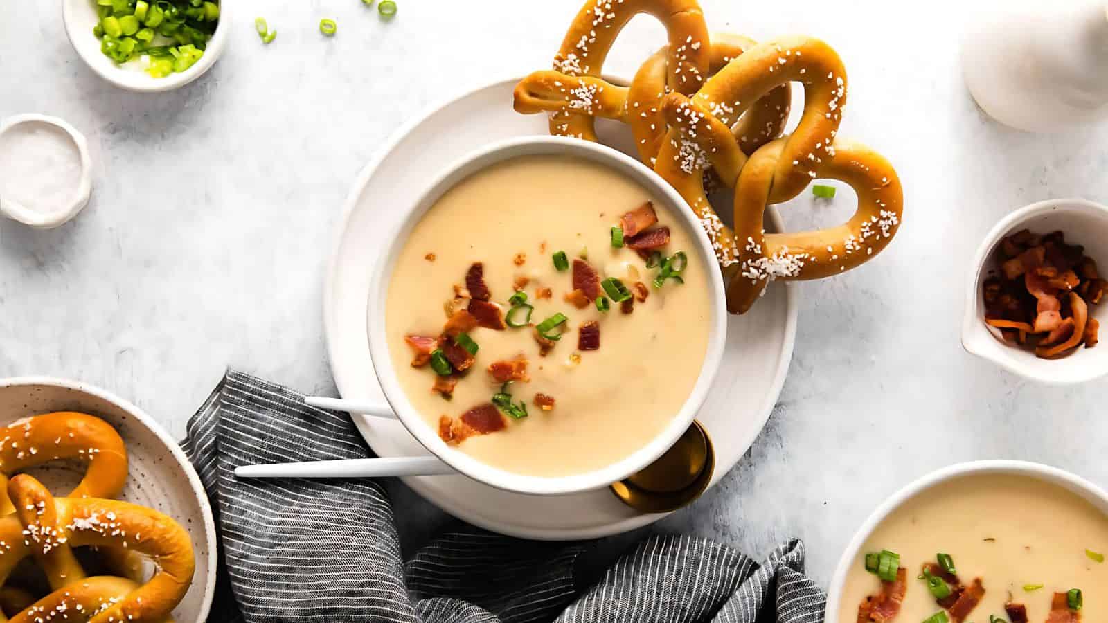 Bowl of creamy soup with bacon and chives, accompanied by soft pretzels on a white table.