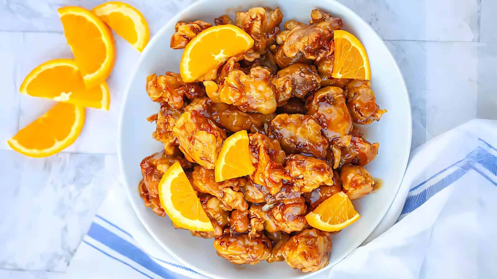A plate of orange chicken garnished with orange slices, placed on a marble surface with additional orange wedges on the side. A striped cloth is partially visible next to the plate.