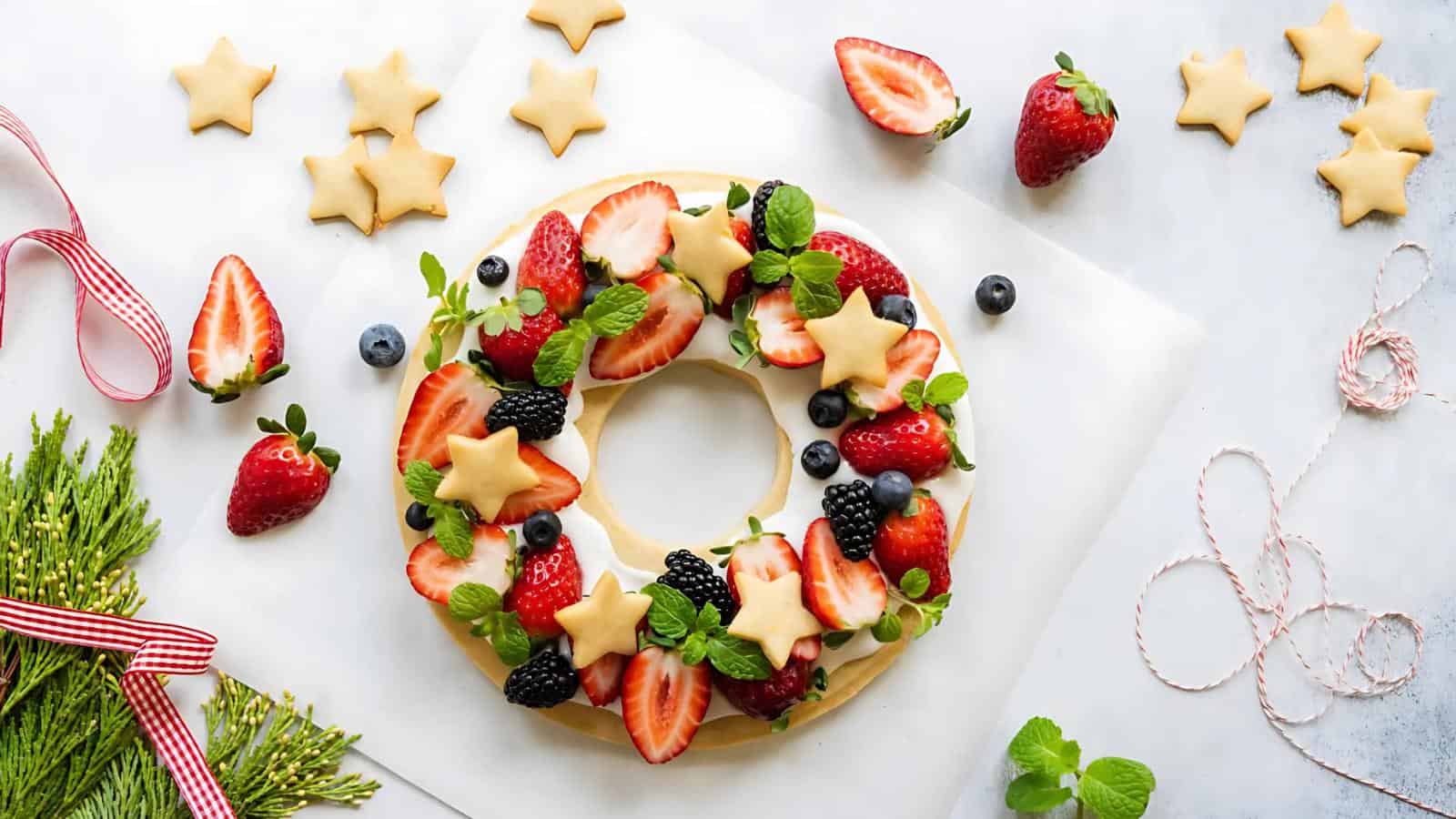 A wreath-shaped dessert decorated with strawberries, blueberries, blackberries, star-shaped cookies, and mint leaves. Surrounded by cookies, ribbons, and greenery on a white surface.