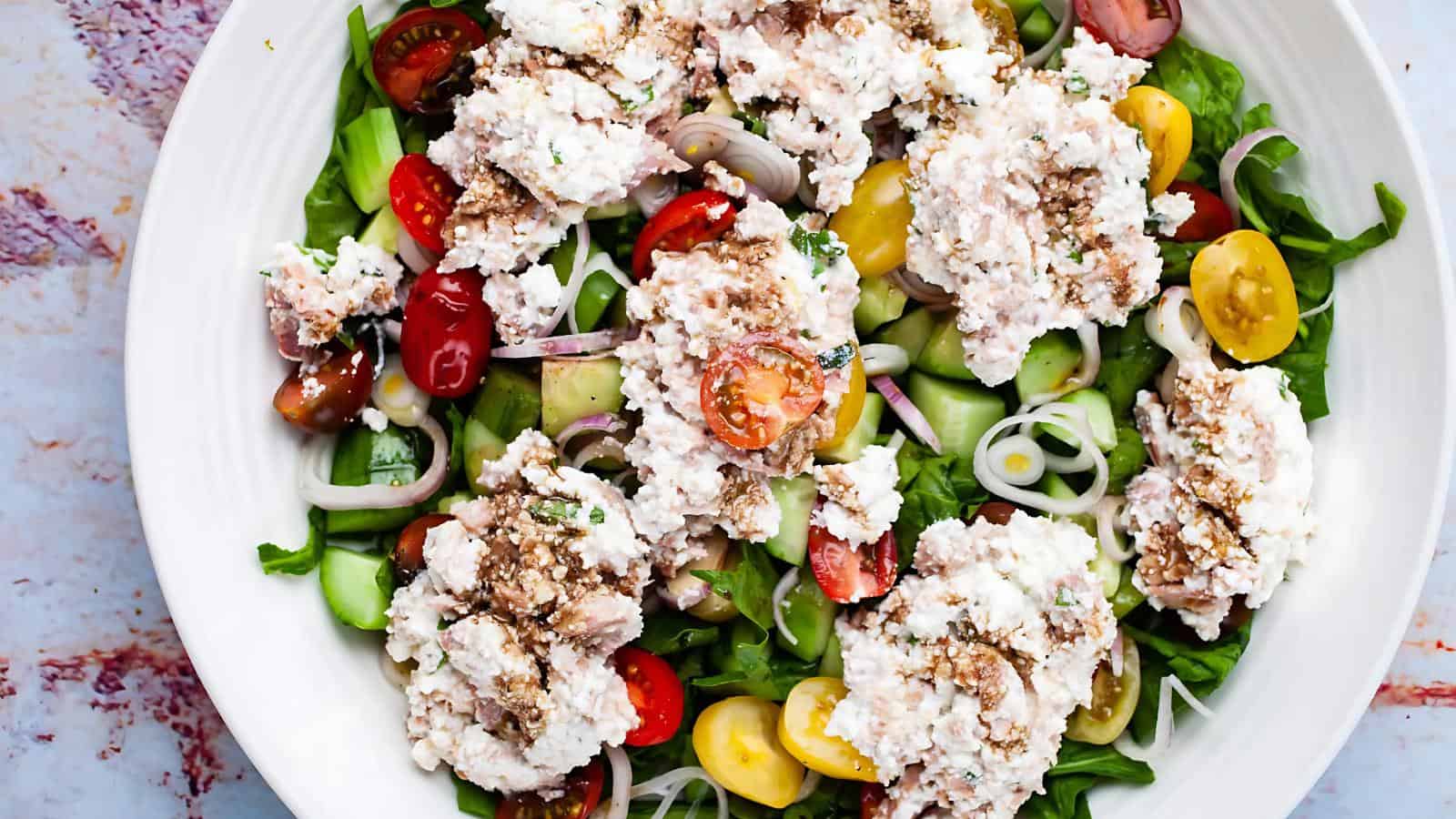 A salad with sliced cucumbers, cherry tomatoes, onions, and greens, topped with dollops of spread and a drizzle of dressing on a white plate.