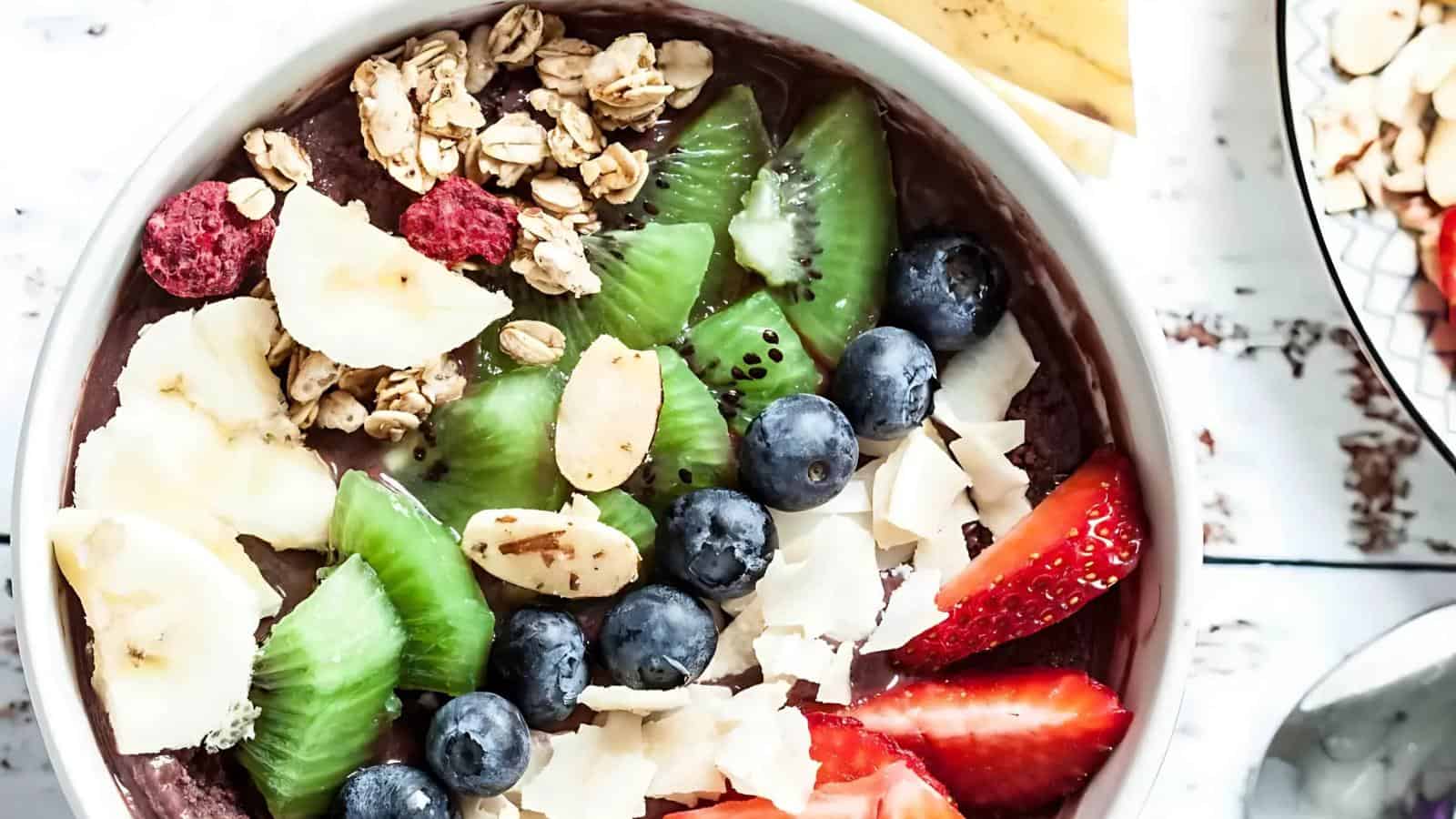 A bowl filled with banana slices, kiwi, blueberries, strawberries, raspberries, granola, coconut flakes, and almonds.