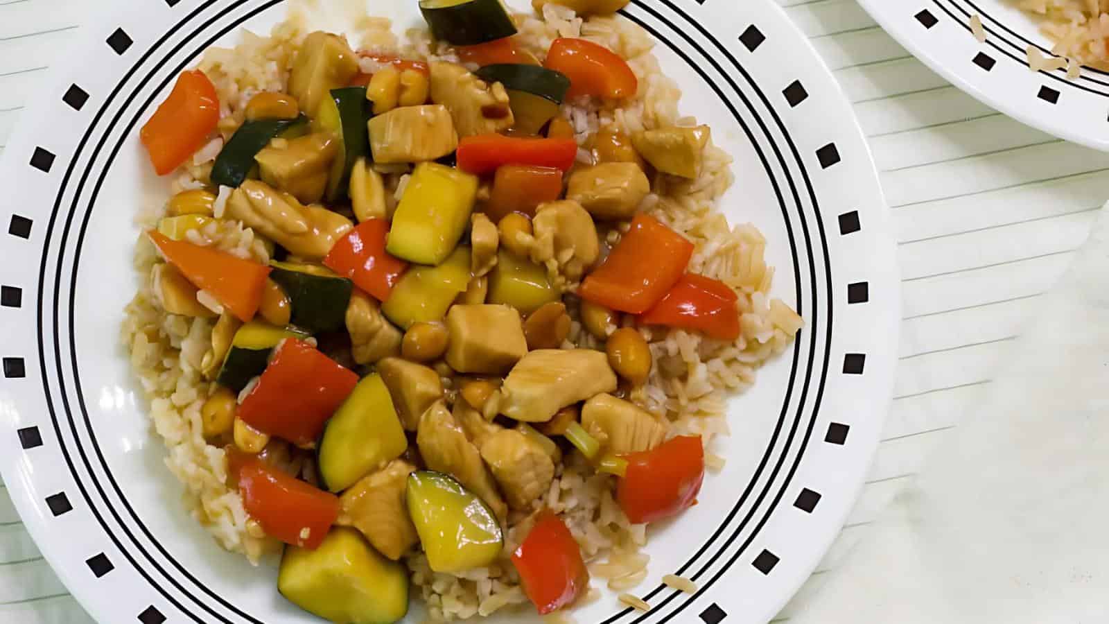 A plate of stir-fried chicken with colorful bell peppers, zucchini, and cashews, served over brown rice on a patterned white plate.