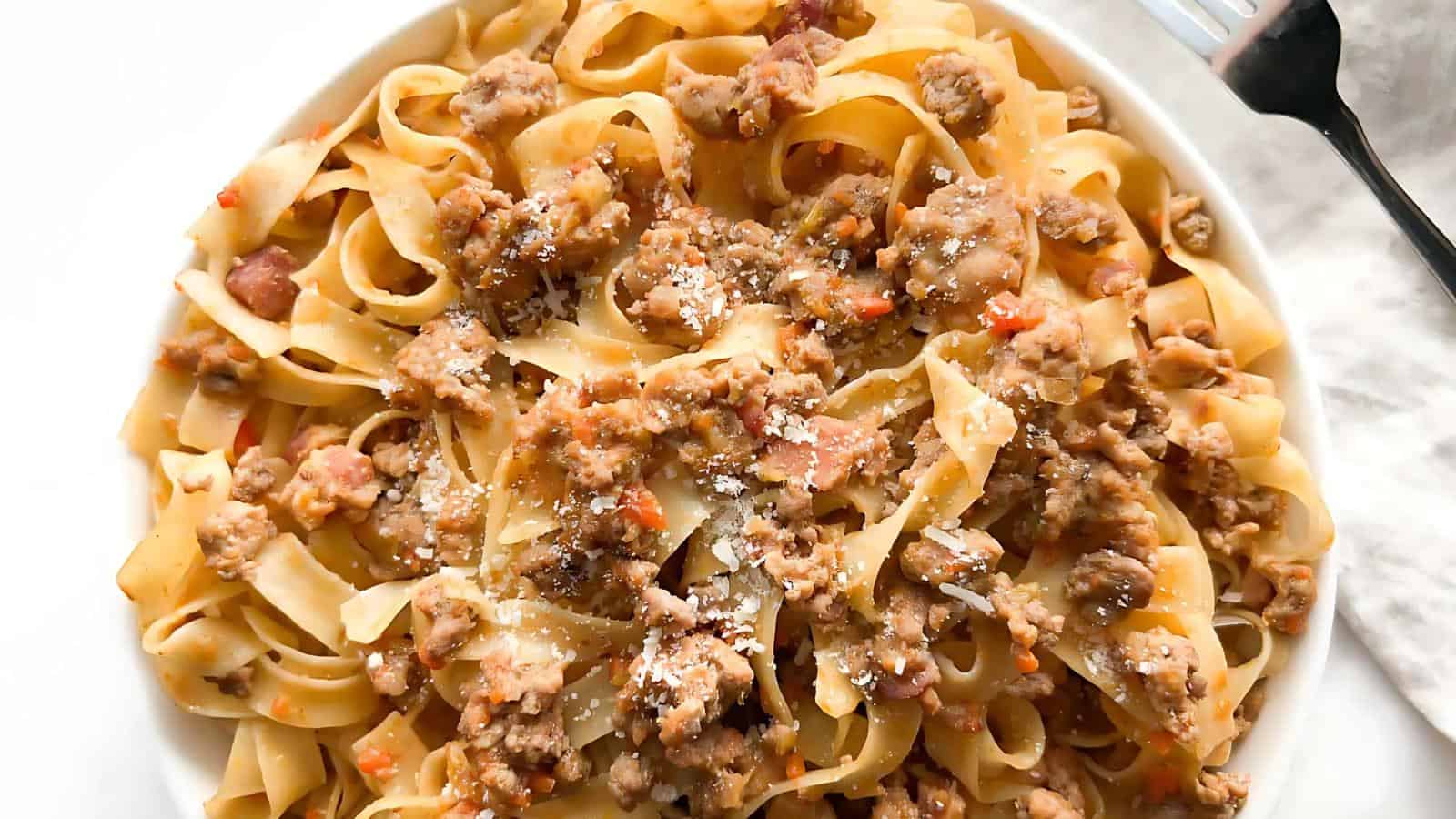A plate of tagliatelle pasta topped with a ground meat sauce and sprinkled with grated cheese, accompanied by a fork on the side.