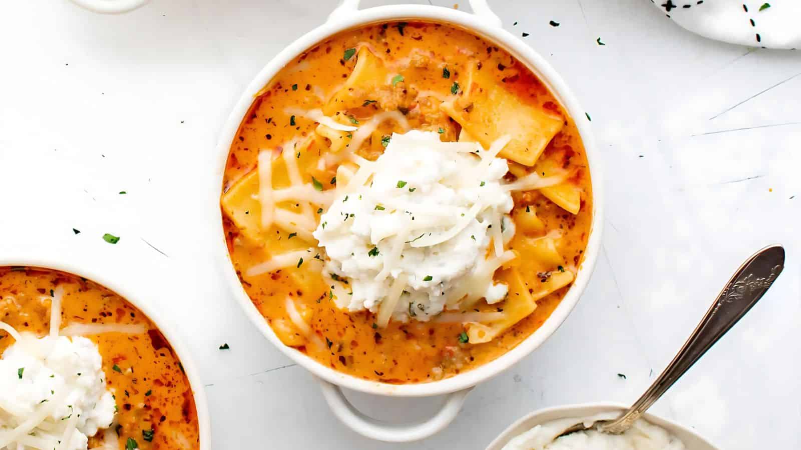 A bowl of creamy lasagna soup topped with shredded cheese and herbs, served in a white bowl with a spoon beside it.