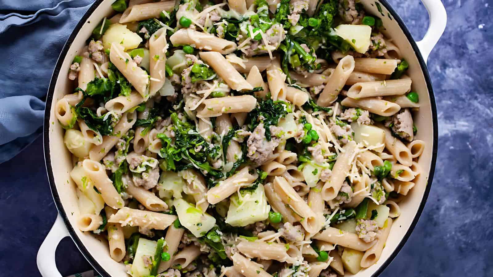 A white skillet filled with penne pasta mixed with greens, peas, and chunks of potato, topped with shredded cheese.