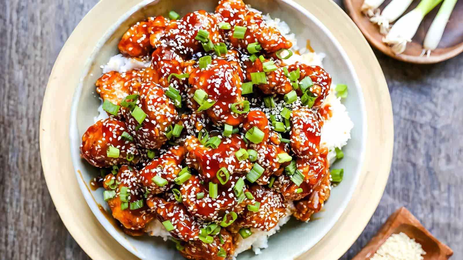 A bowl of rice topped with glazed chicken pieces, garnished with chopped green onions and sesame seeds.