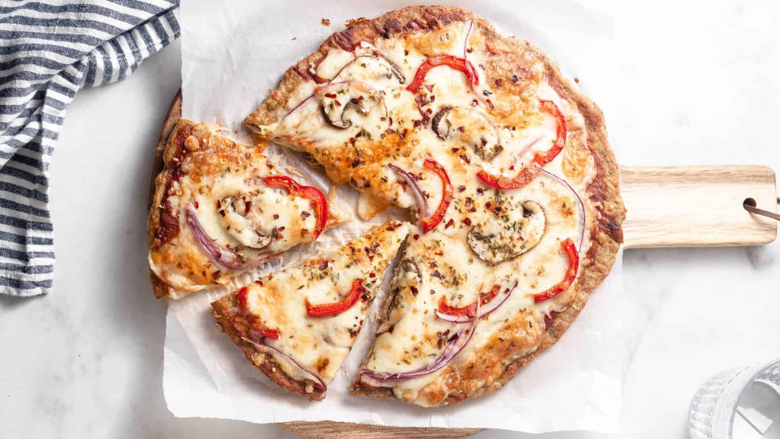 A baked pizza with melted cheese, red bell pepper slices, mushrooms, and red onion is topped with chili flakes on a wooden cutting board. Perfect for those exploring low carb lunch recipes, it sits elegantly beside a striped cloth.