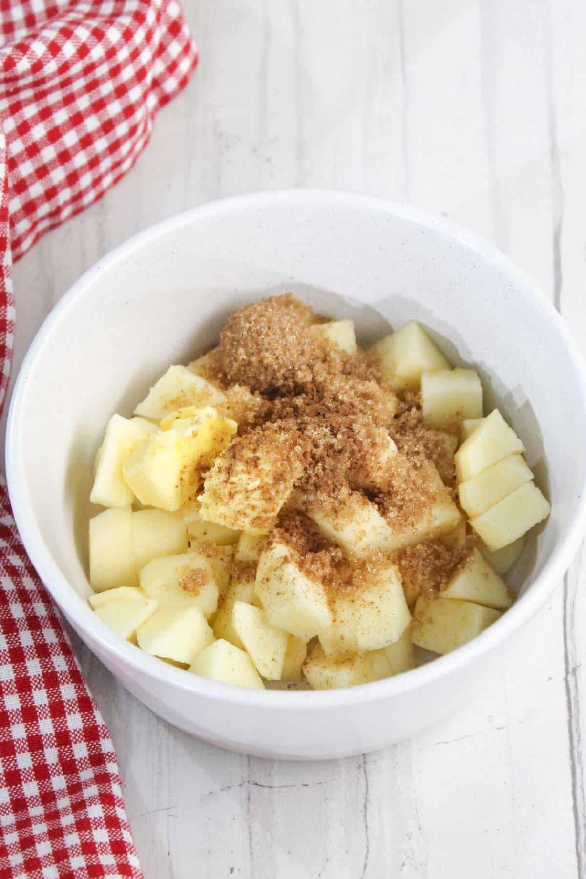 A white bowl containing diced apples mixed with brown sugar, cinnamon, and other spices on a white surface, next to a red and white checkered cloth.