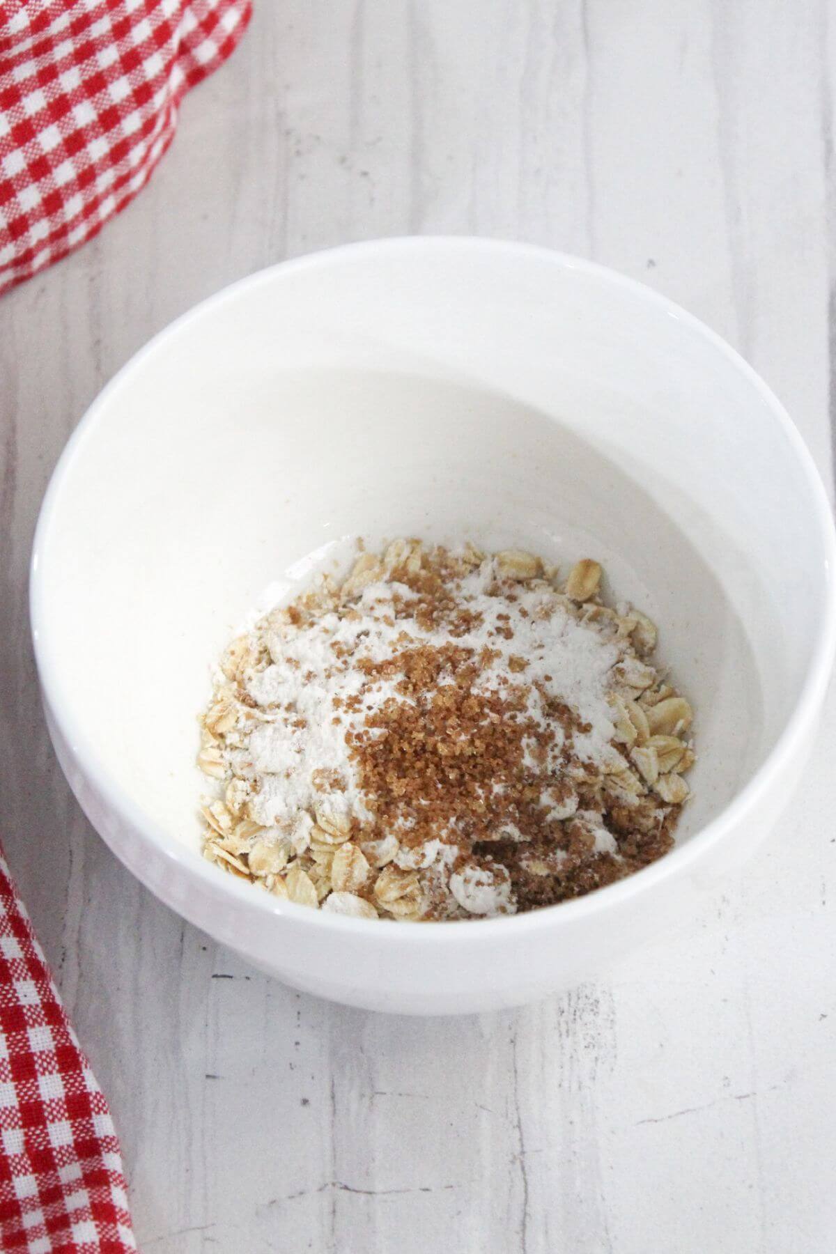 A white bowl containing oats, flour, and brown sugar on a white countertop next to a red and white checkered cloth.