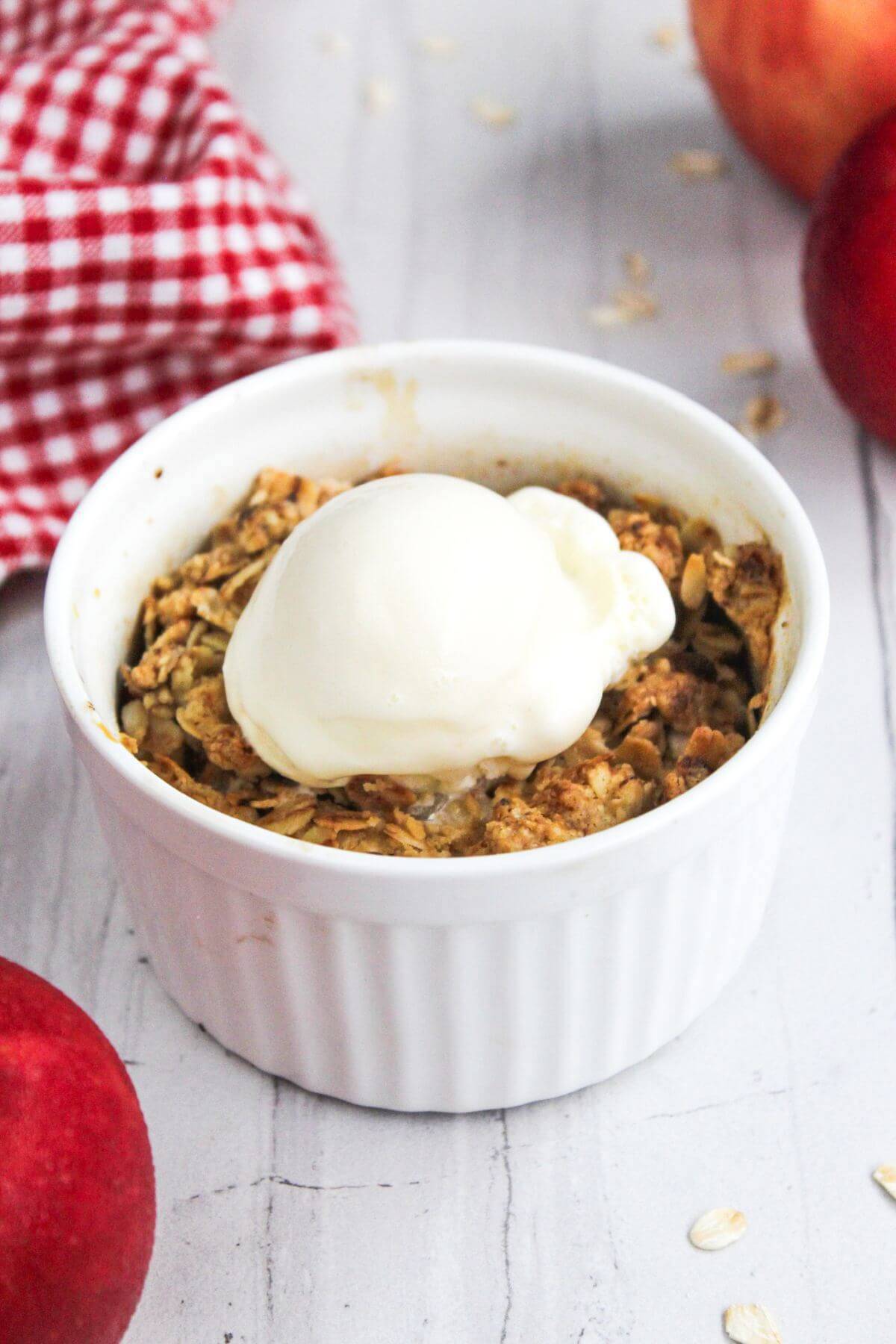 A white ramekin containing a baked dessert topped with a scoop of vanilla ice cream, surrounded by fresh apples and a red and white checkered cloth.