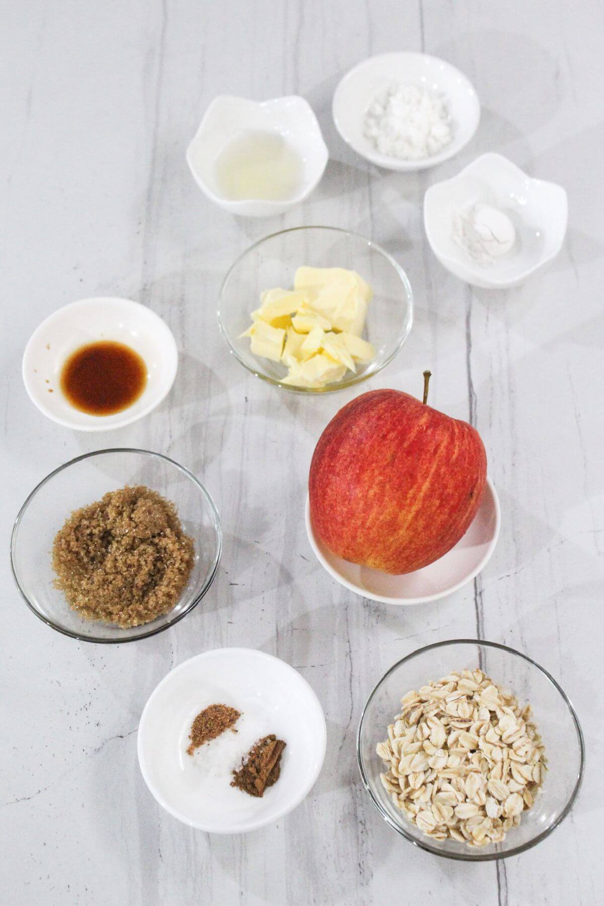 An array of ingredients including an apple, butter, vanilla extract, brown sugar, oats, spices, and small bowls of various powders, arranged on a white surface.