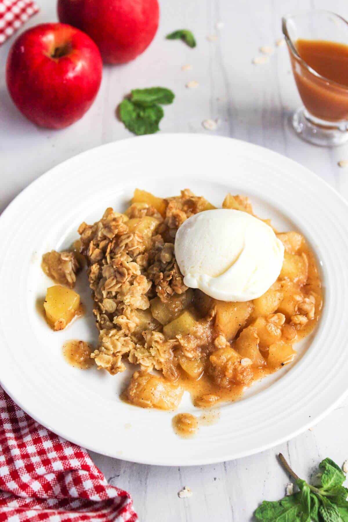 A plate of apple crisp topped with a scoop of vanilla ice cream. Surrounding the plate are two apples, a small pitcher of caramel sauce, and a red and white checkered cloth.