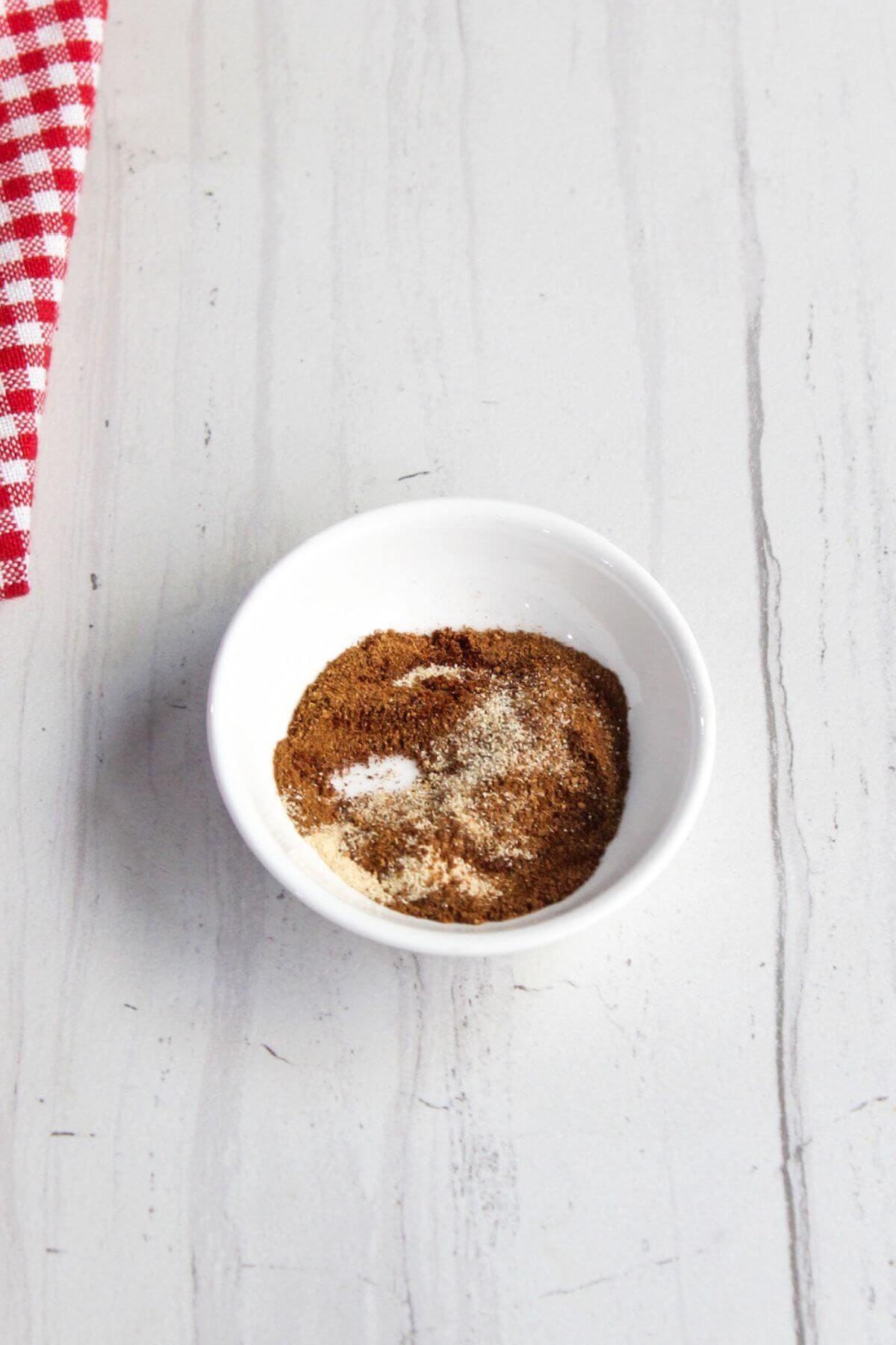 A small white bowl with brown seasoning on a white marble surface next to a red and white checkered cloth.