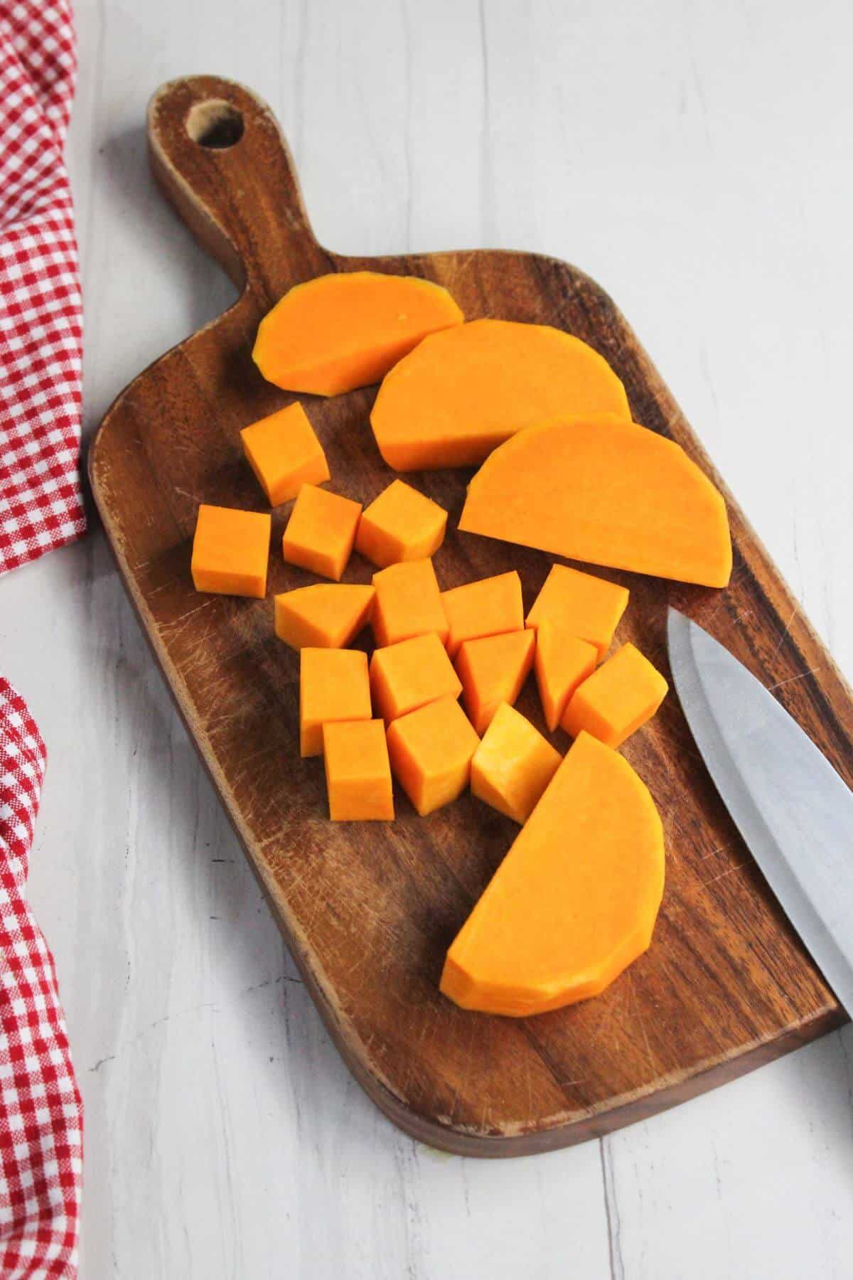 Cubed and sliced butternut squash on a wooden cutting board with a knife next to a red and white checkered cloth.