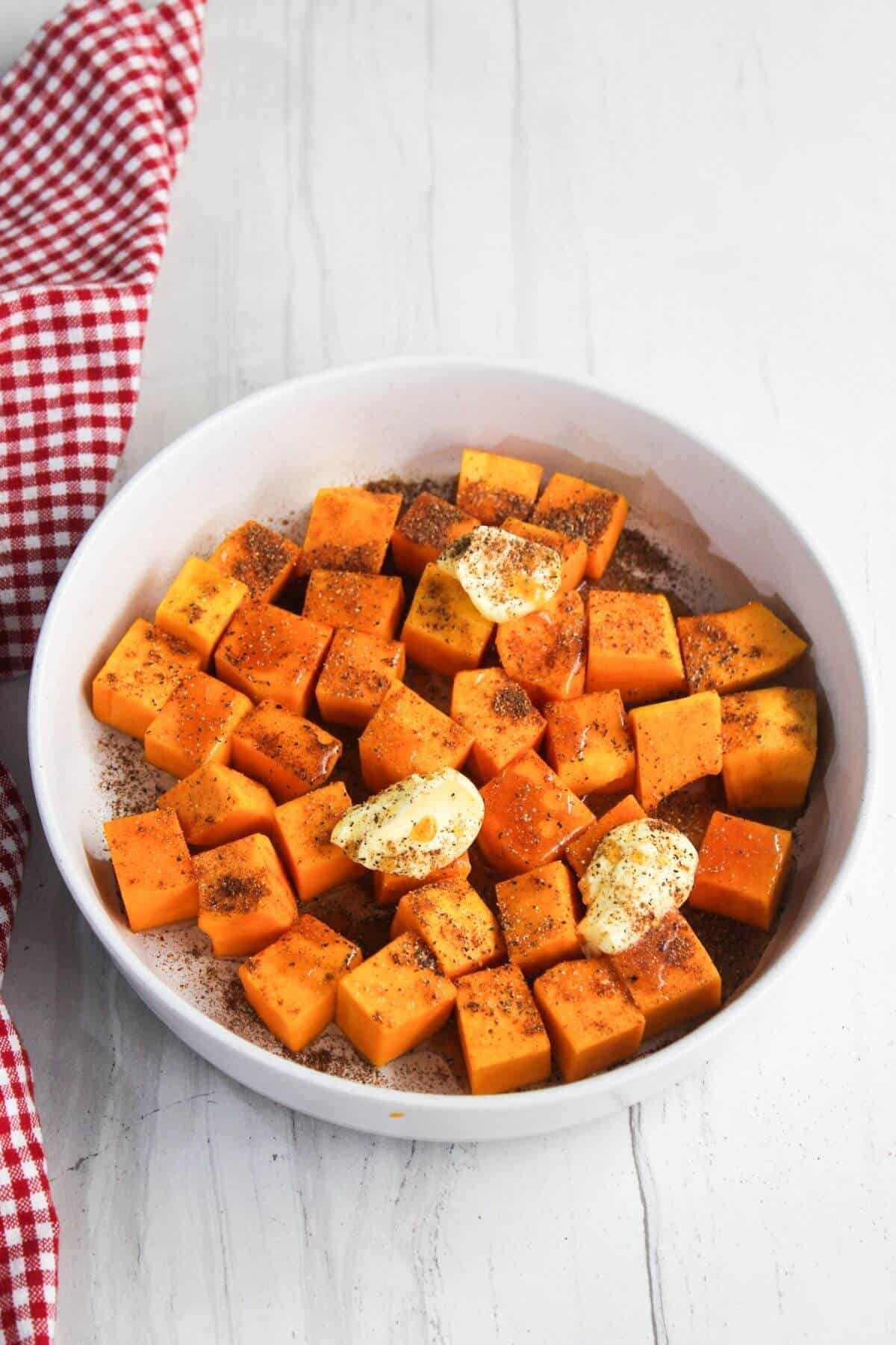 White bowl with cubed butternut squash, topped with butter and spices. A red and white checkered cloth is partially visible on the left side.