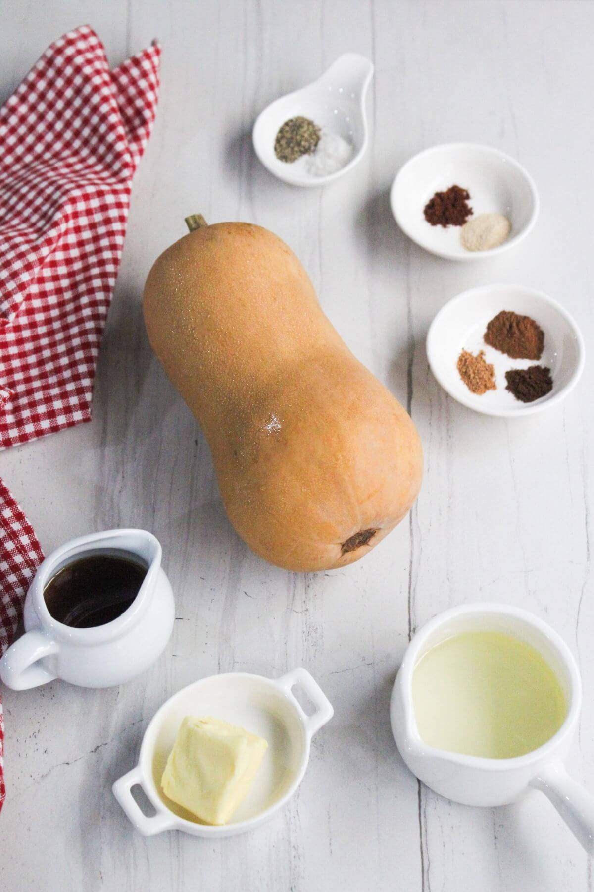 A whole butternut squash surrounded by small dishes containing spices, butter, oil, and syrup on a white surface with a red checkered cloth.