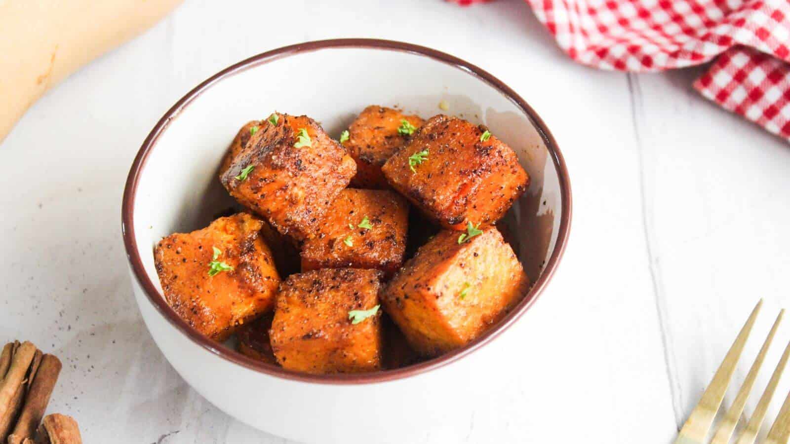 A bowl of roasted sweet potato cubes garnished with herbs, placed on a white surface. A red and white checkered cloth is partially visible in the background.