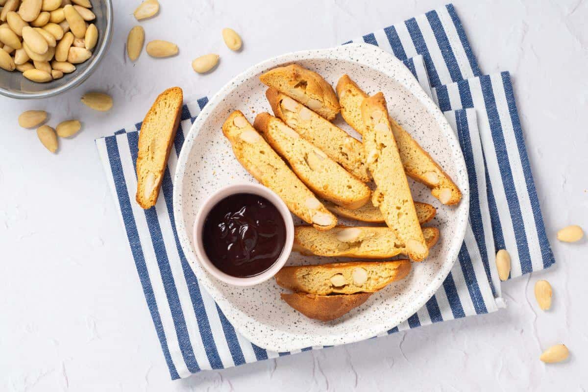 A plate of biscotti with almonds is served with a small bowl of dark dipping sauce. A small bowl of almonds is nearby on a striped cloth.