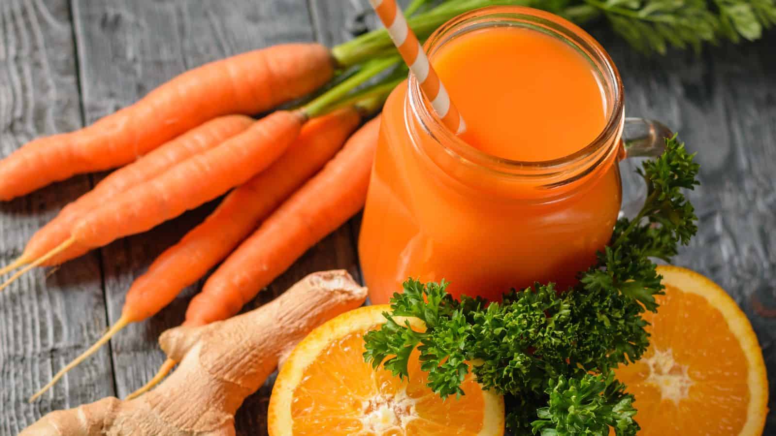 A jar of carrot juice with a striped straw, surrounded by fresh carrots, ginger, orange slices, and parsley on a wooden surface.