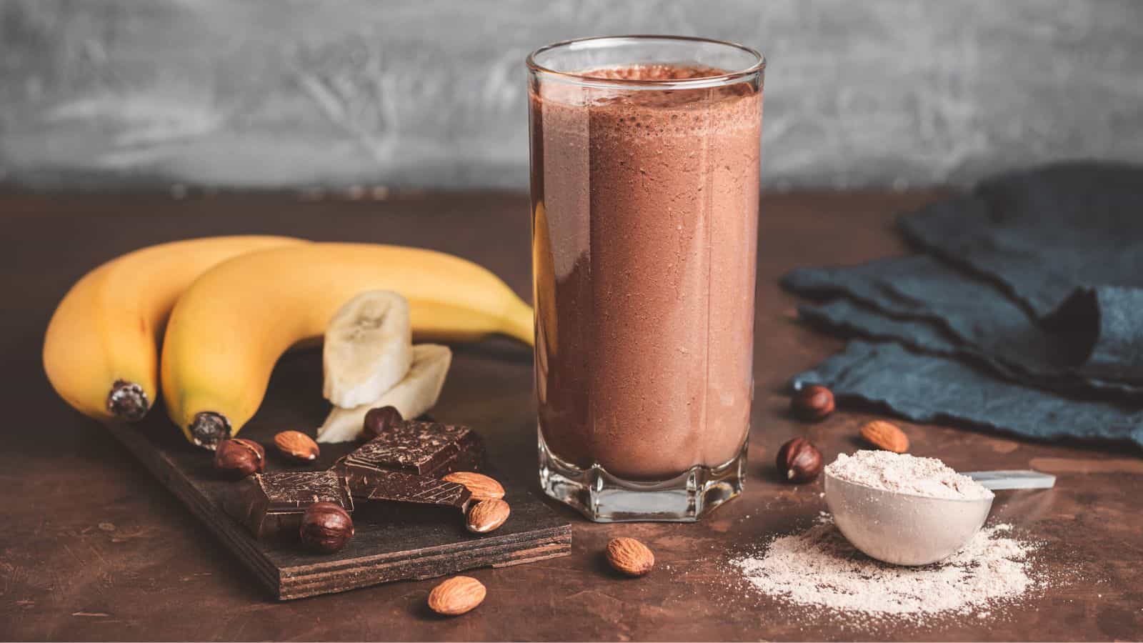 A glass of chocolate smoothie is on a table with bananas, chocolate, hazelnuts, almonds, and a scoop of powder beside it.