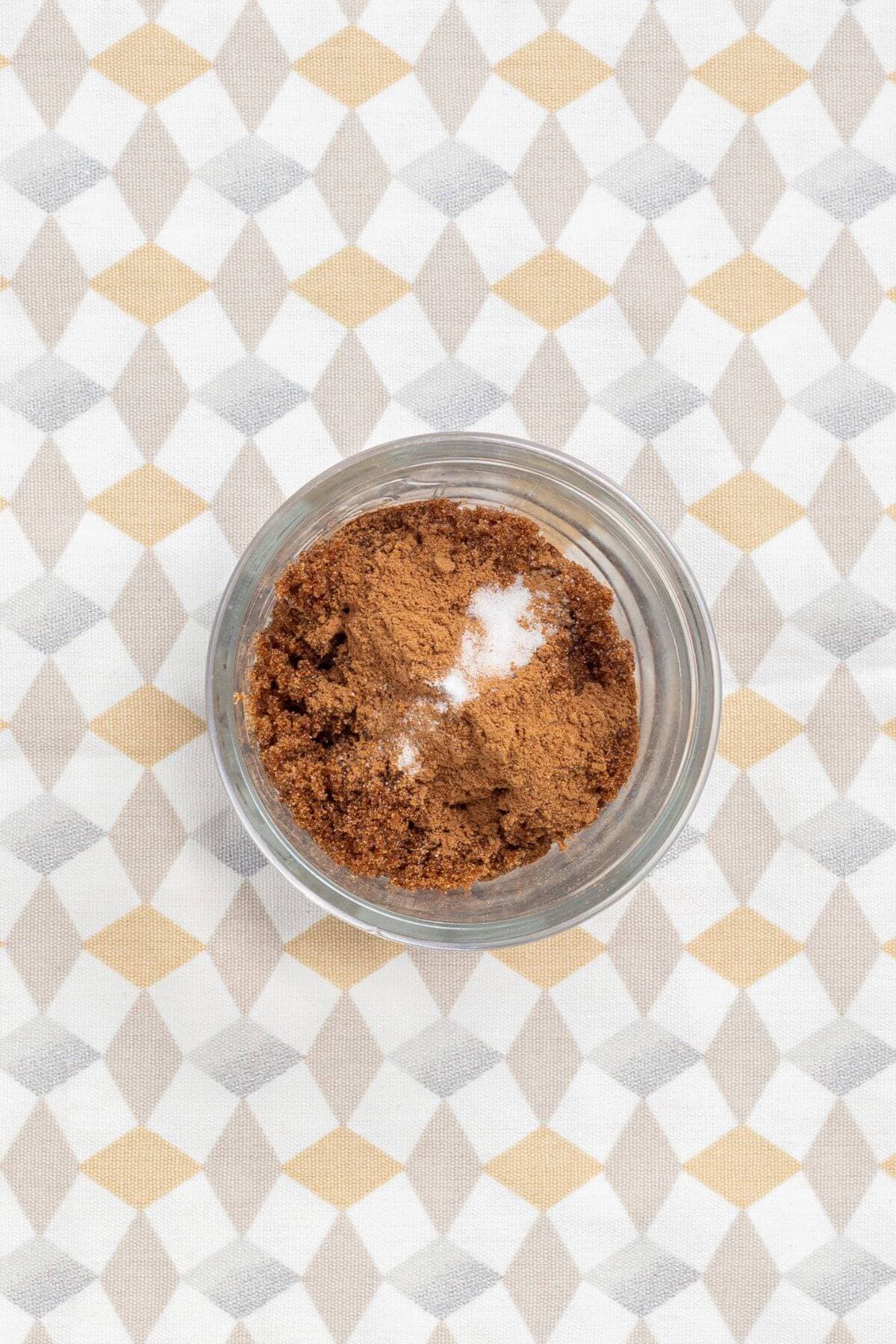 A glass bowl containing a mixture of brown sugar and white powder, possibly baking soda, on a geometric patterned surface.