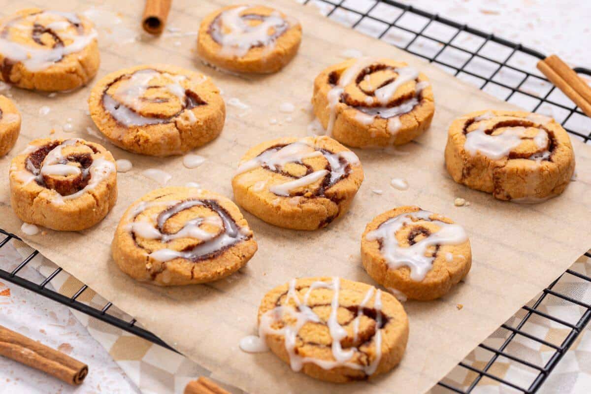 A batch of glazed cinnamon roll cookies on a parchment-lined cooling rack, with cinnamon sticks scattered around.
