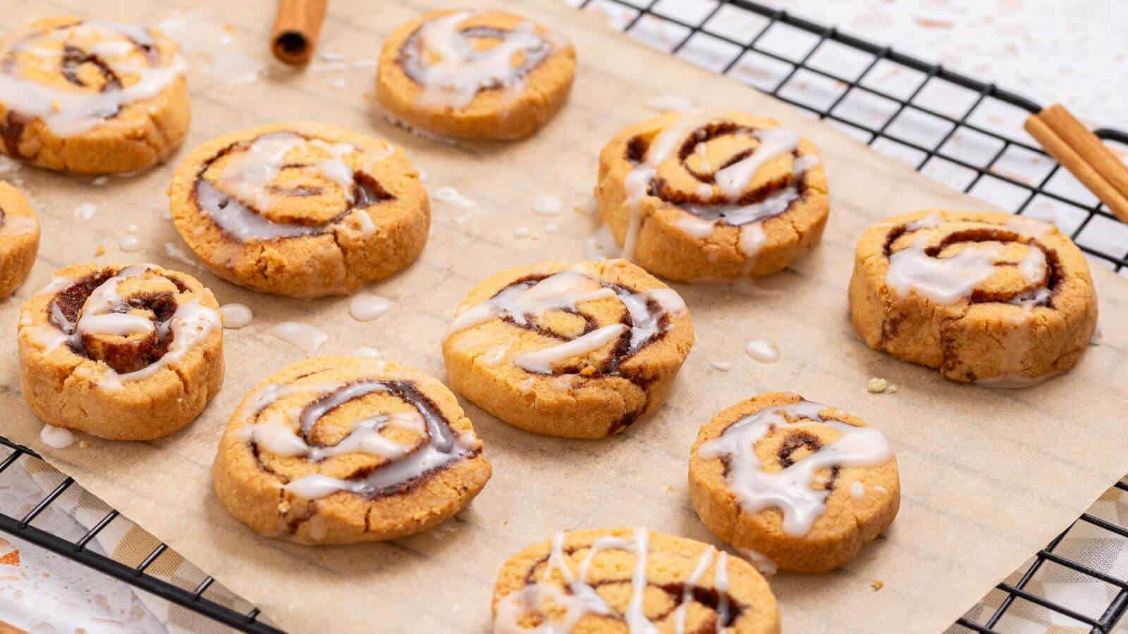 Cinnamon swirl cookies with icing on a parchment-lined cooling rack, with cinnamon sticks nearby.