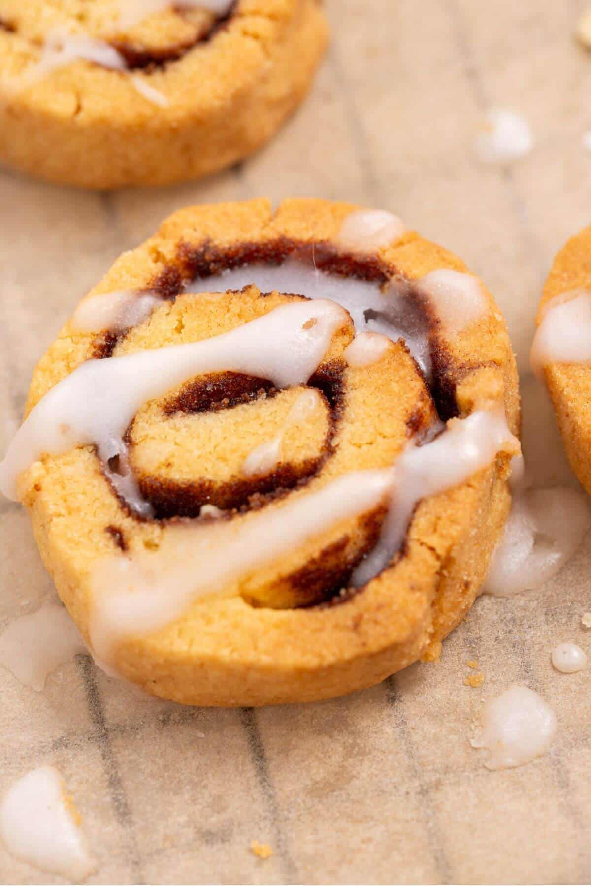 Close-up of a cinnamon roll cookie topped with white icing, placed on parchment paper.