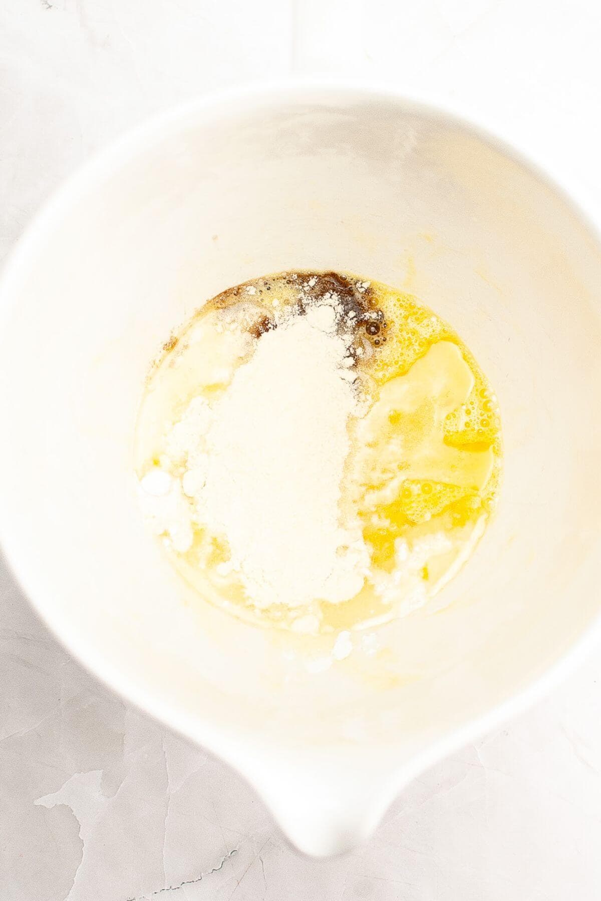 A mixing bowl containing flour, melted butter, and brown sugar on a white surface.