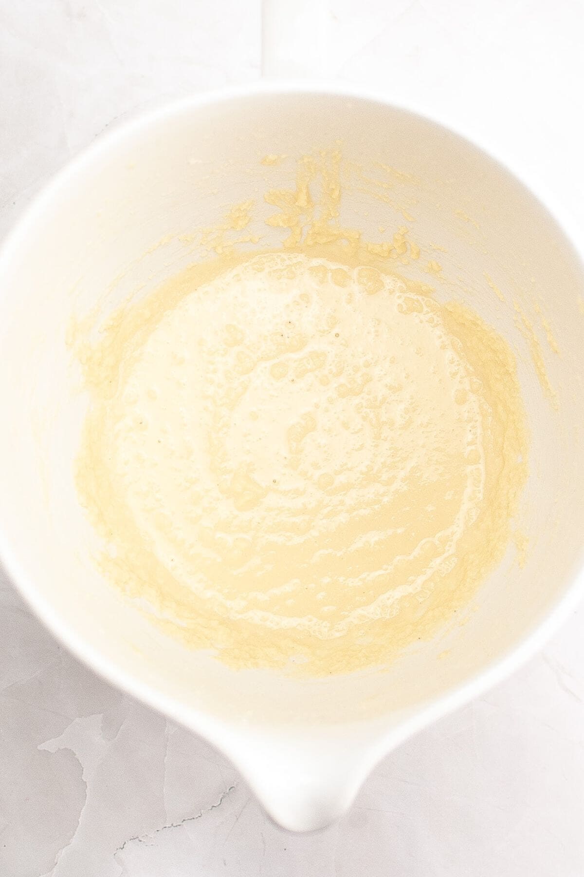 A white mixing bowl contains a creamy, pale yellow batter on a white surface.