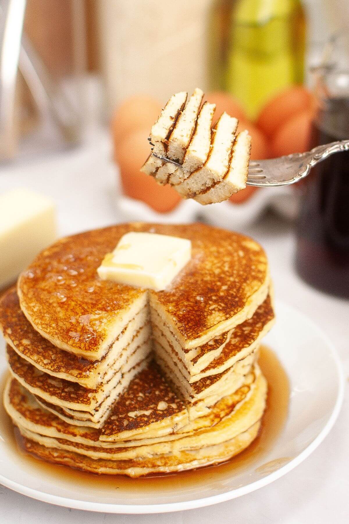 Stack of pancakes topped with a pat of butter on a plate, surrounded by syrup. A fork holds a cut piece above the stack.