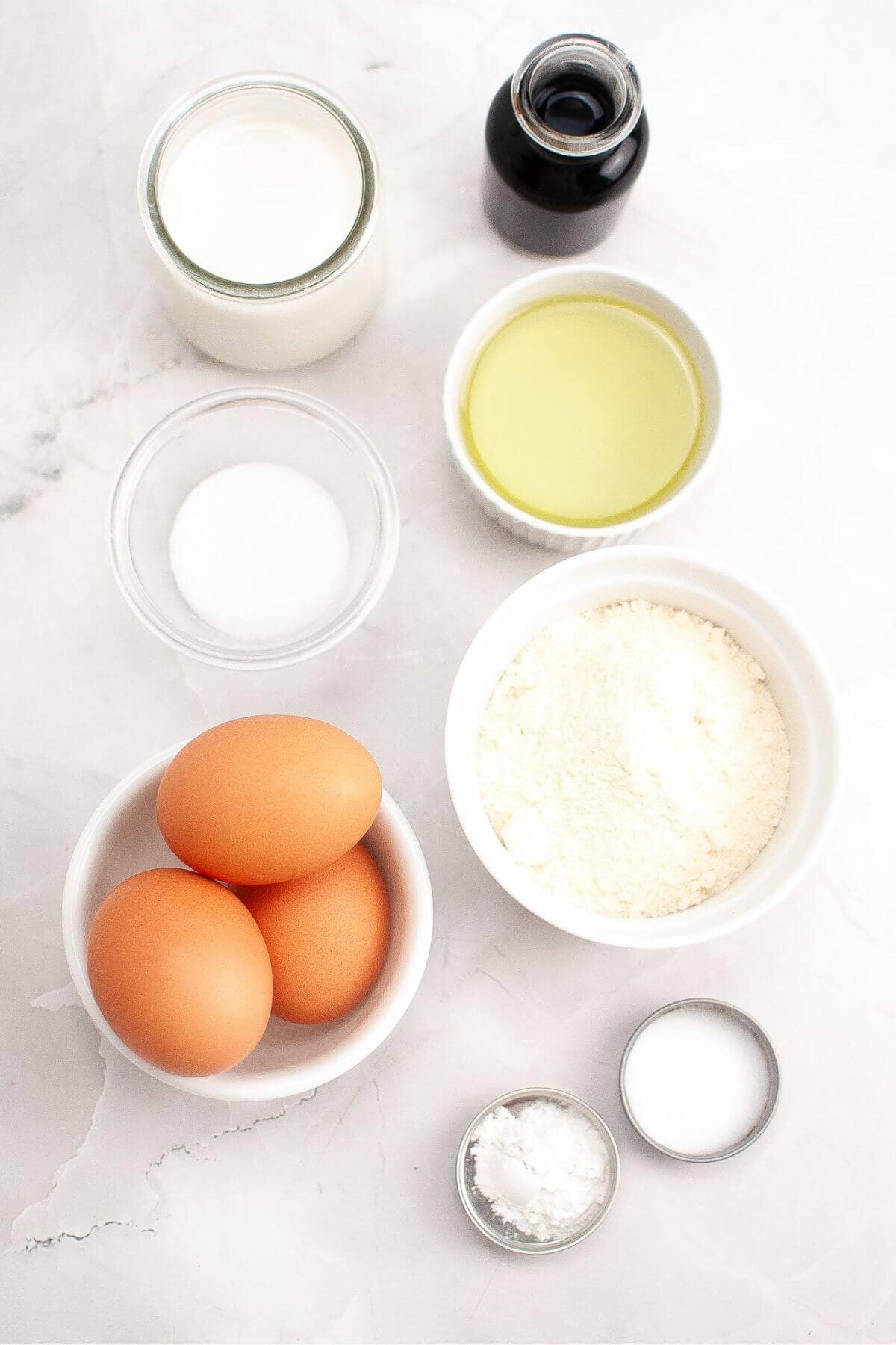 Ingredients for baking on a marble surface, including eggs, flour, oil, milk, vanilla extract, sugar, baking powder, and salt.