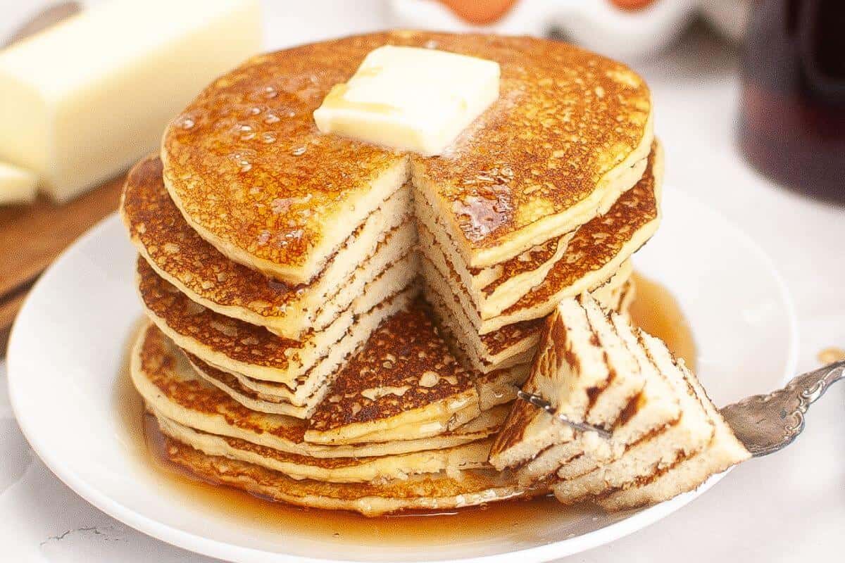 A stack of pancakes with a pat of butter on top, served on a white plate with syrup, and a fork holding a cut piece.