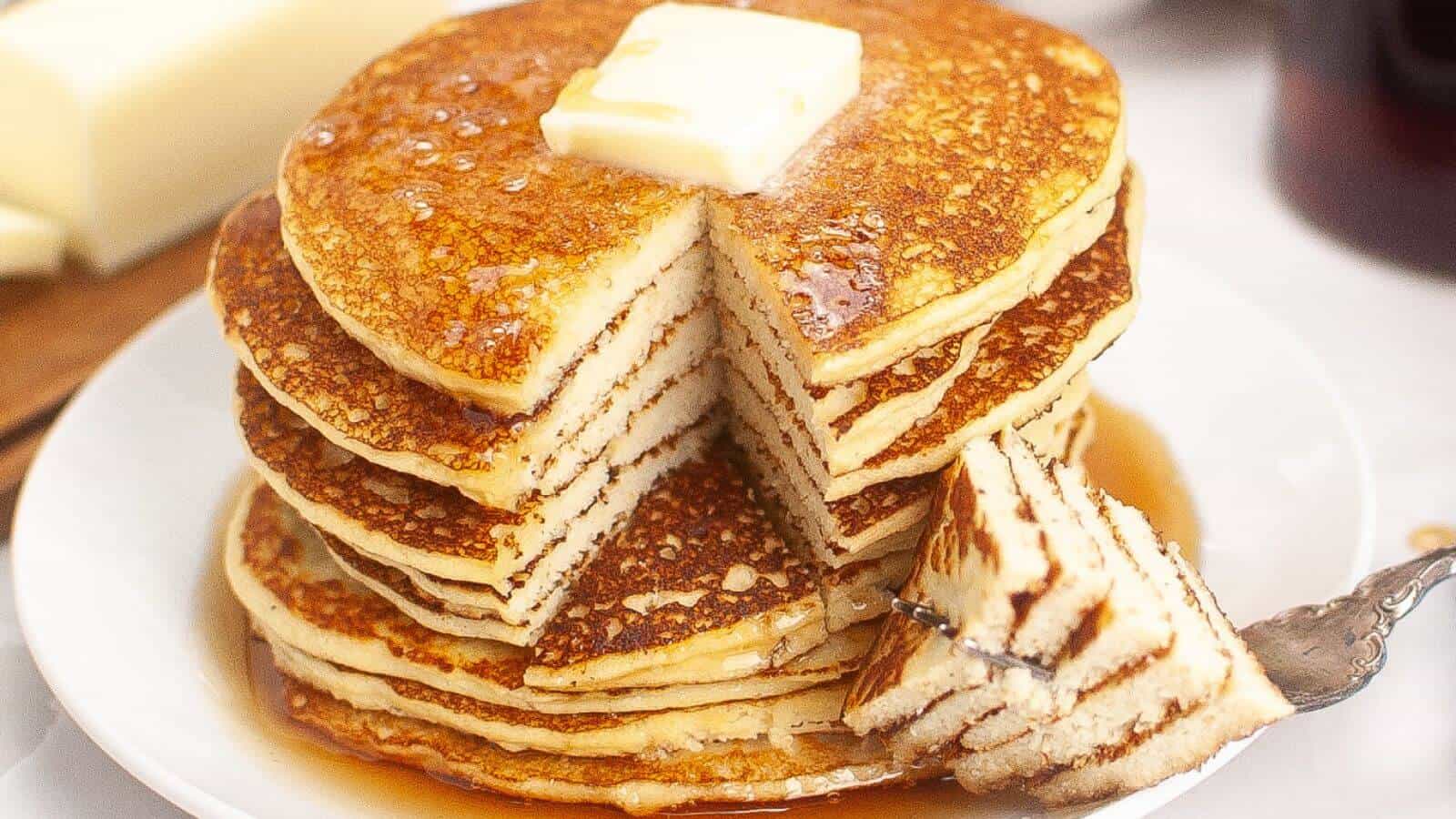 A stack of pancakes with a pat of butter on top, drizzled with syrup. A portion is cut out and placed on a fork, resting on the plate.