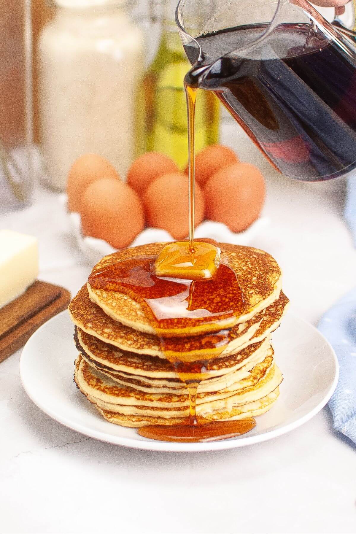 Syrup being poured over a stack of pancakes on a white plate. Eggs and kitchen ingredients are in the background.
