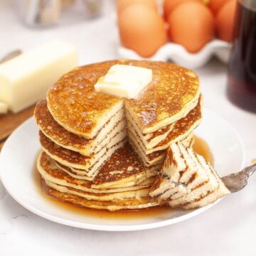 A stack of pancakes with a slice cut out, topped with a butter pat and syrup, on a white plate. Eggs, butter, and syrup bottle in the background.
