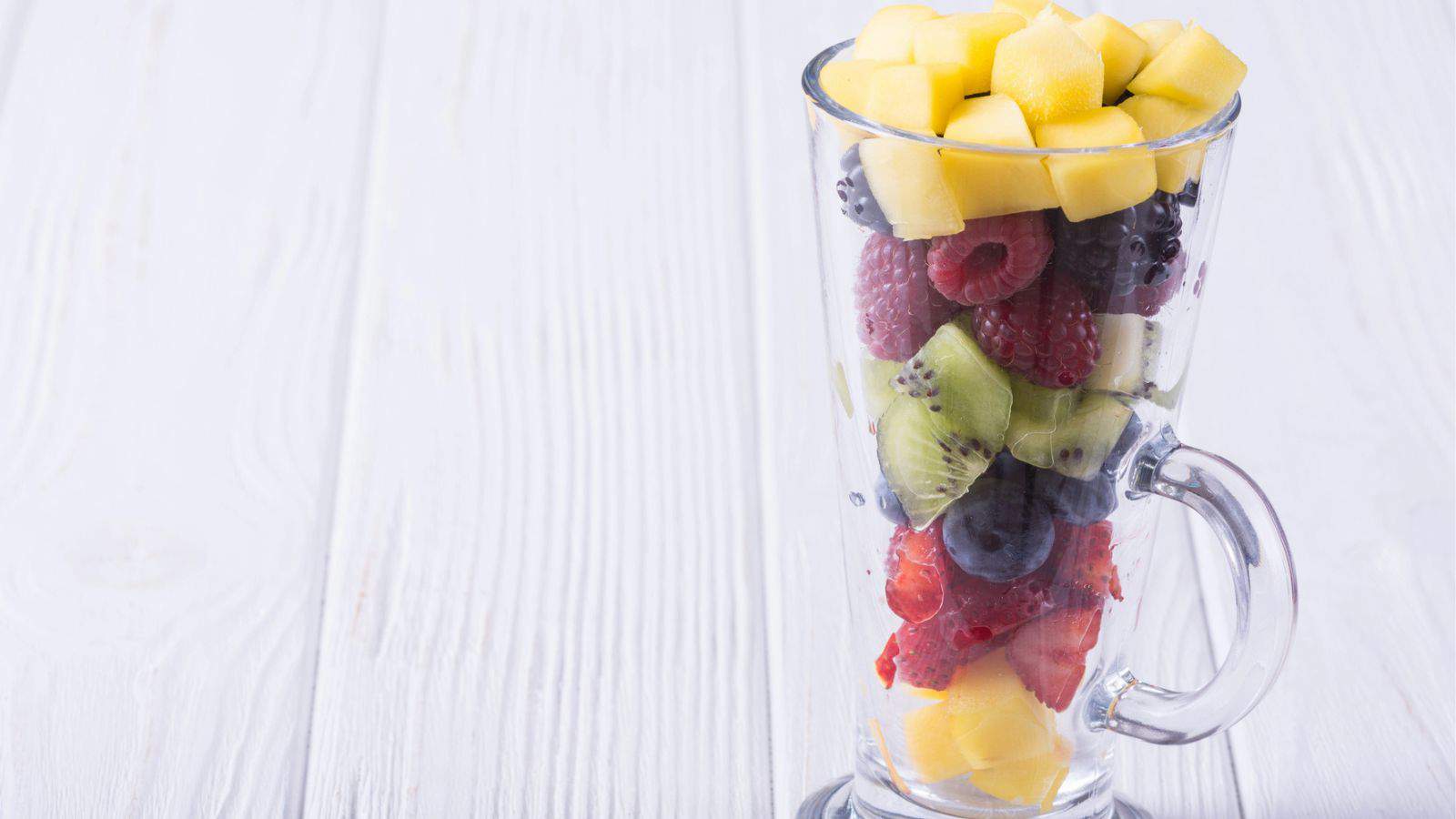 A tall glass filled with layers of diced mango, raspberries, blackberries, kiwi slices, blueberries, and strawberries on a white wooden surface.