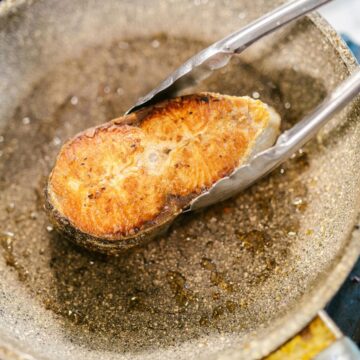 A piece of salmon is being seared in a frying pan, held by tongs over a stove.