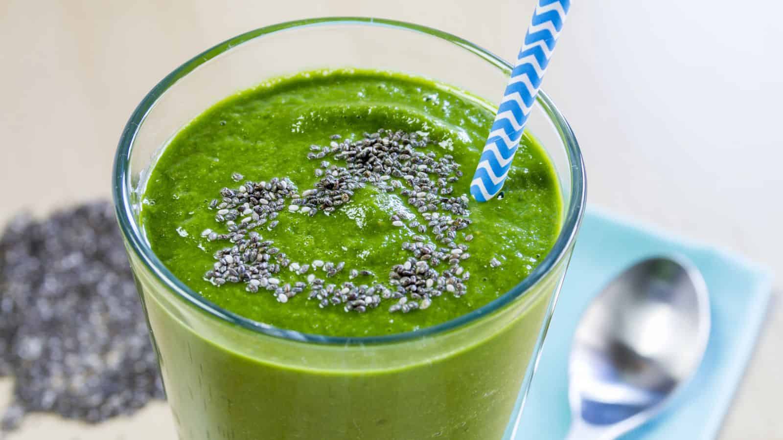 A glass of green smoothie topped with chia seeds, with a striped straw. A spoon and additional chia seeds are visible in the background.