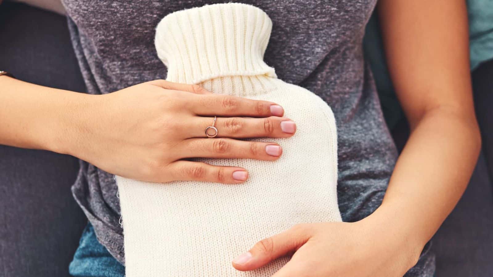 A person wearing a grey shirt holds a cream-colored hot water bottle against their abdomen. Their nails are neatly manicured and they wear a simple ring.
