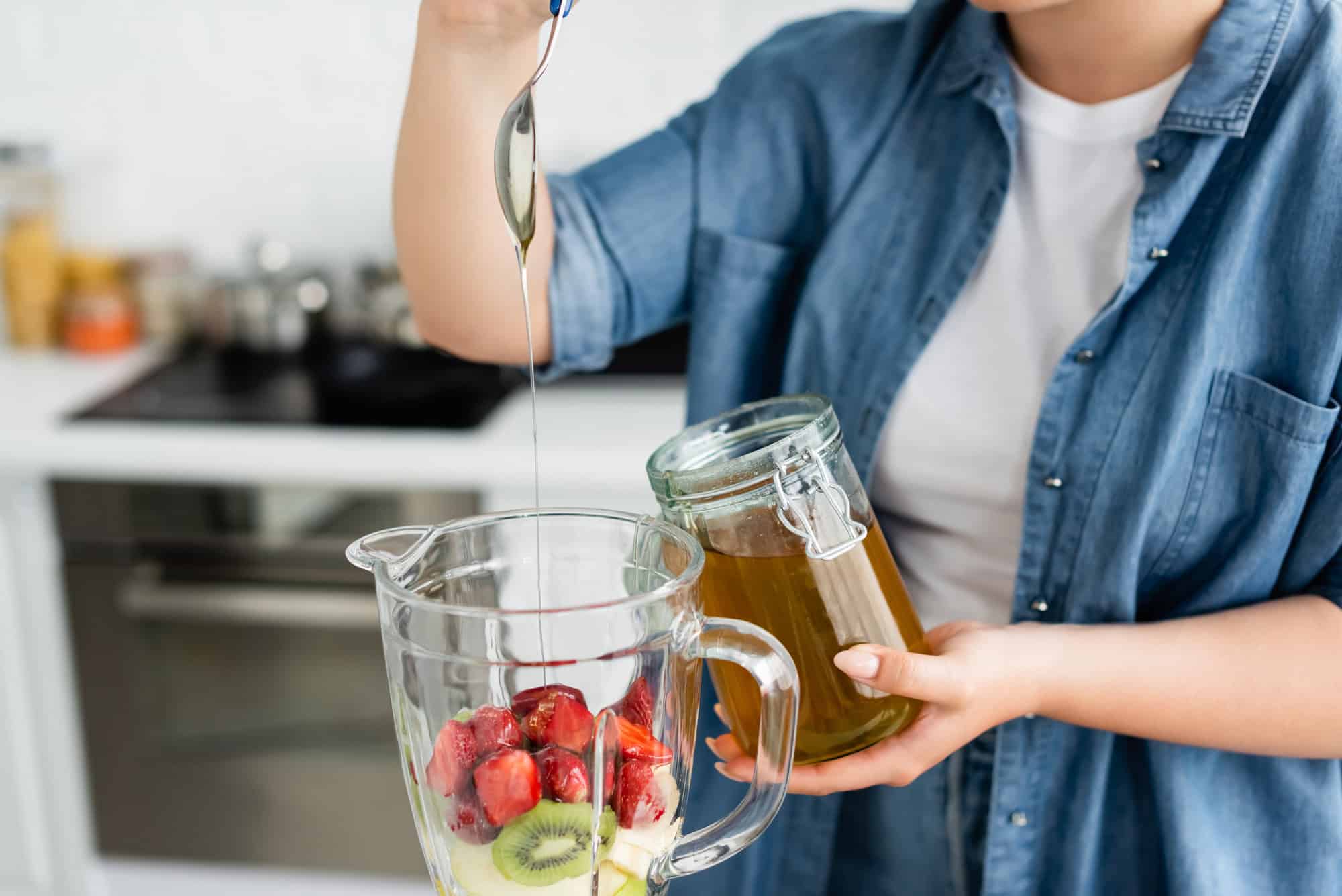 Person pouring honey into a blender containing strawberries and kiwi slices.