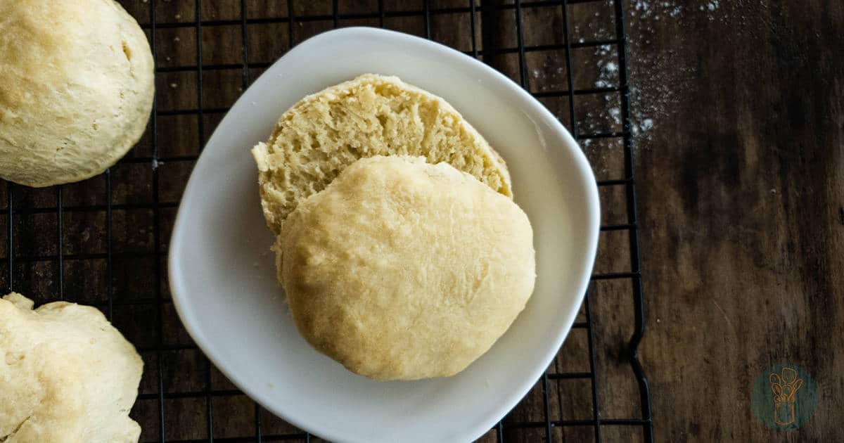 A biscuit, reminiscent of those knockoff recipes so good you won't miss the real thing, is split open on a white plate placed on a cooling rack with additional biscuits nearby.