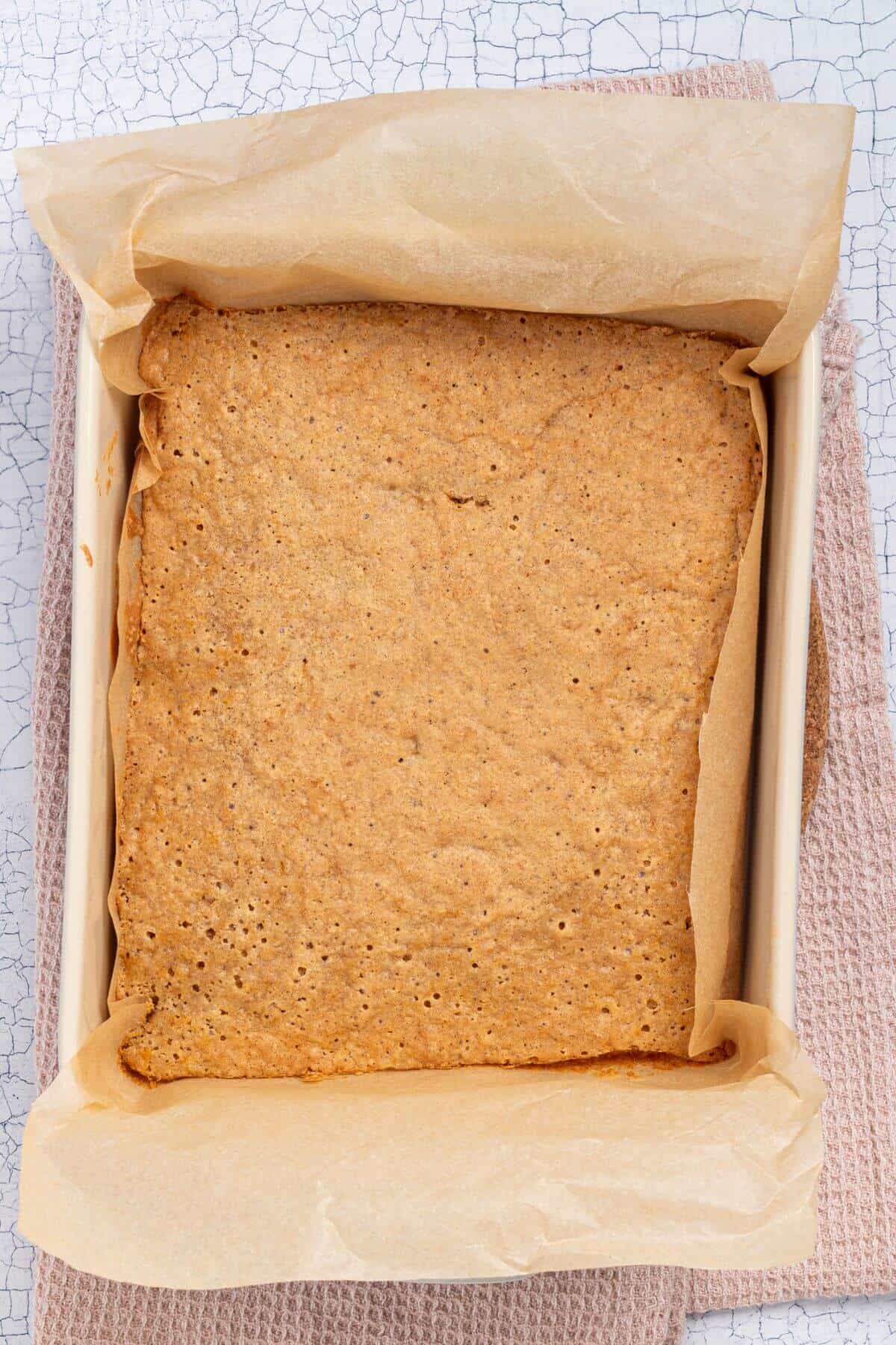 A rectangular, golden-brown baked cake in a parchment-lined baking dish, resting on a textured surface.