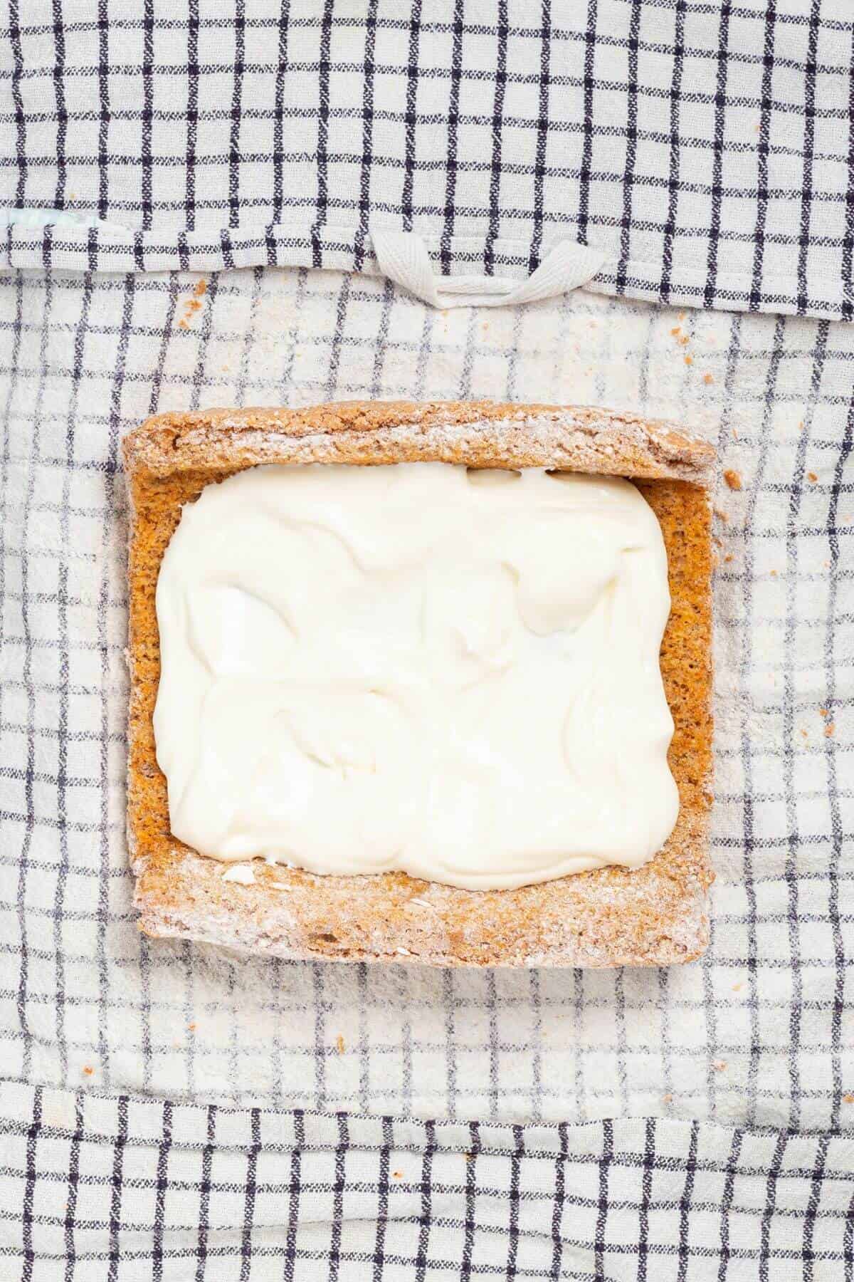 A slice of brown bread with a thick, even layer of cream cheese placed on a white and black checkered cloth.
