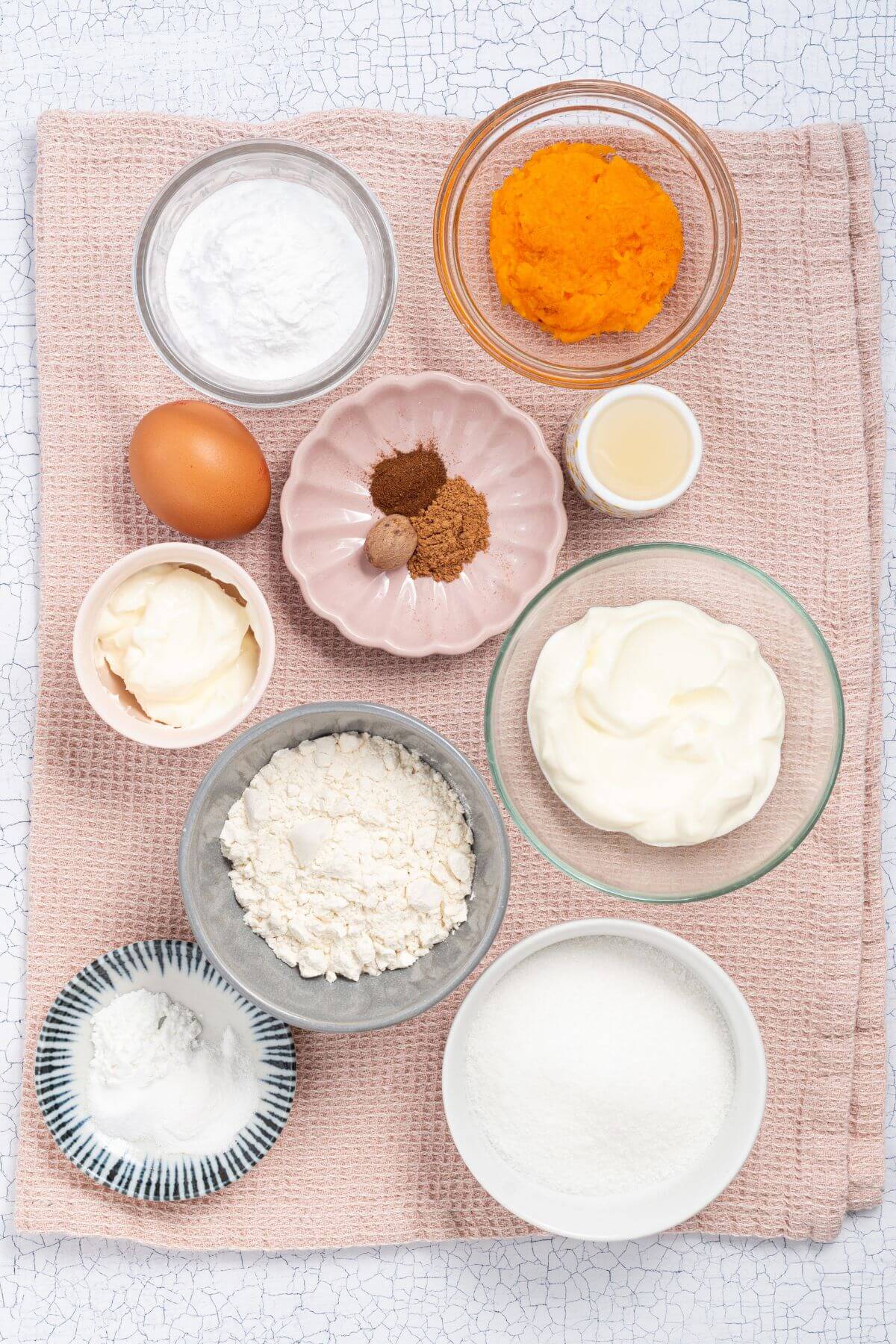 An overhead view of various baking ingredients on a textured pink cloth, including flour, sugar, an egg, spices, baking powder, baking soda, yogurt, pumpkin puree, vanilla extract, and butter.
